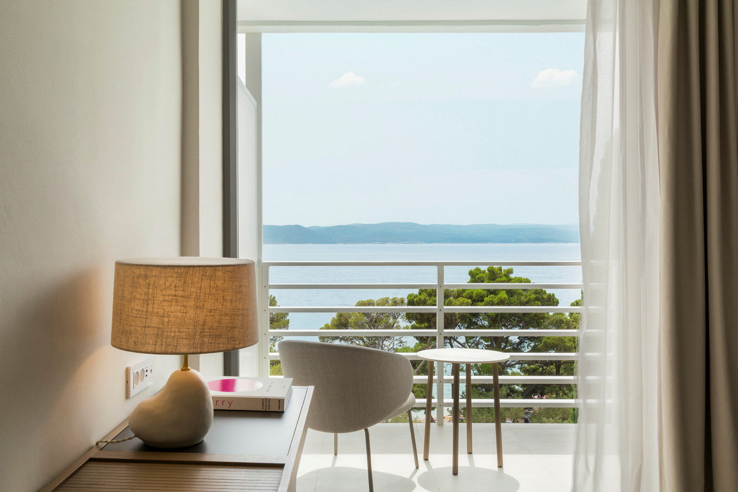 Bilde av et rom med en lampe på et bord, en stol og et lite bord som står ved en balkong med utsikt over havet. (AI-generert tekst)