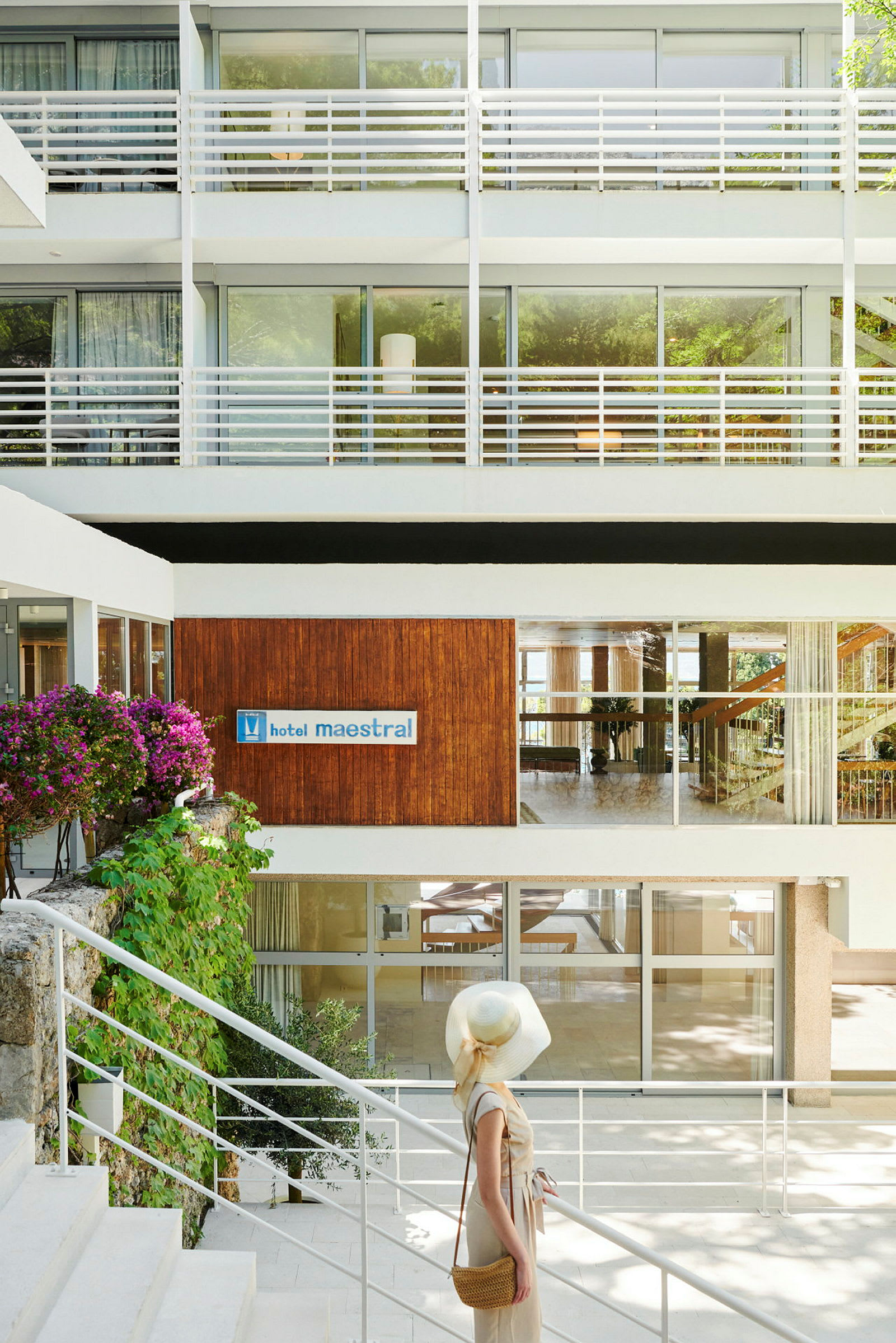 En kvinne står ved en trapp foran hotellets inngang, omgitt av moderne bygningselementer og blomster. (AI-generert tekst)