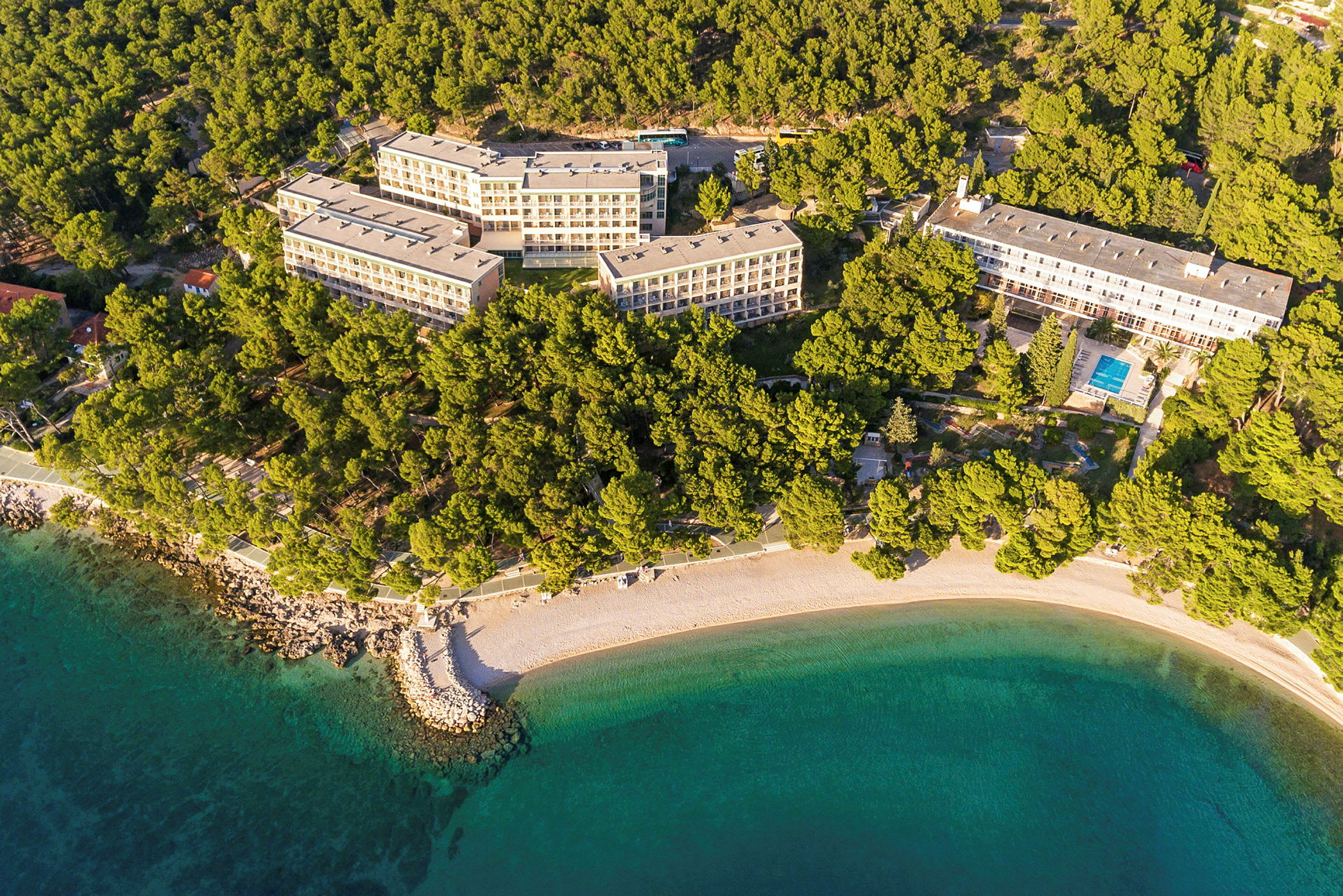Flyfoto av hotellkompleks omgitt av skog og strand. (AI-generert tekst)