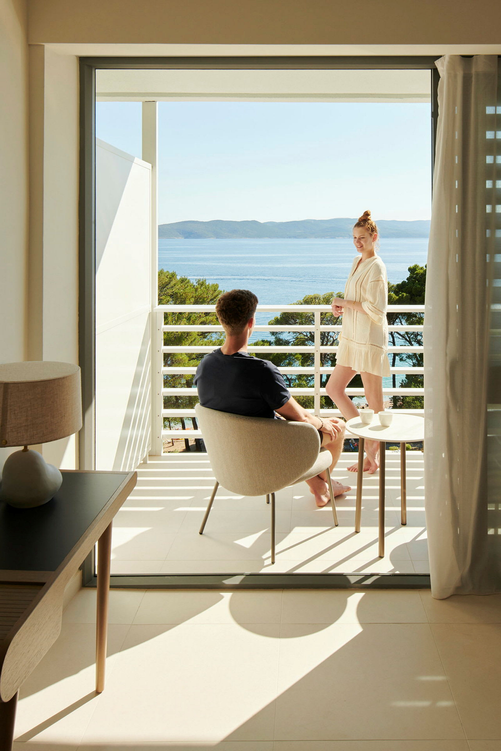En hotellbalkong med utsikt mot havet og omkringliggende natur. En person sitter og en annen står ved siden av bordet. (AI-generert tekst)