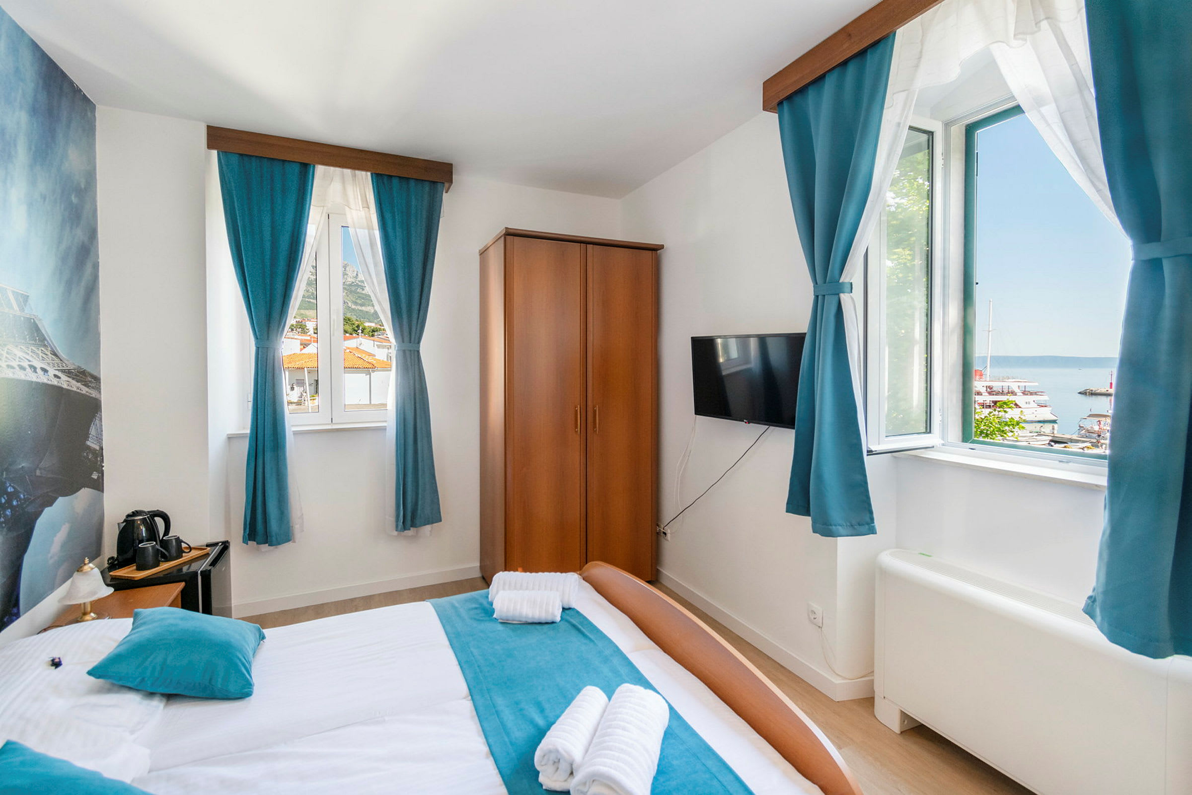 Hotellrom med dobbeltseng, blå gardiner og utsikt over en havn utenfor vinduet. (AI-generert tekst)