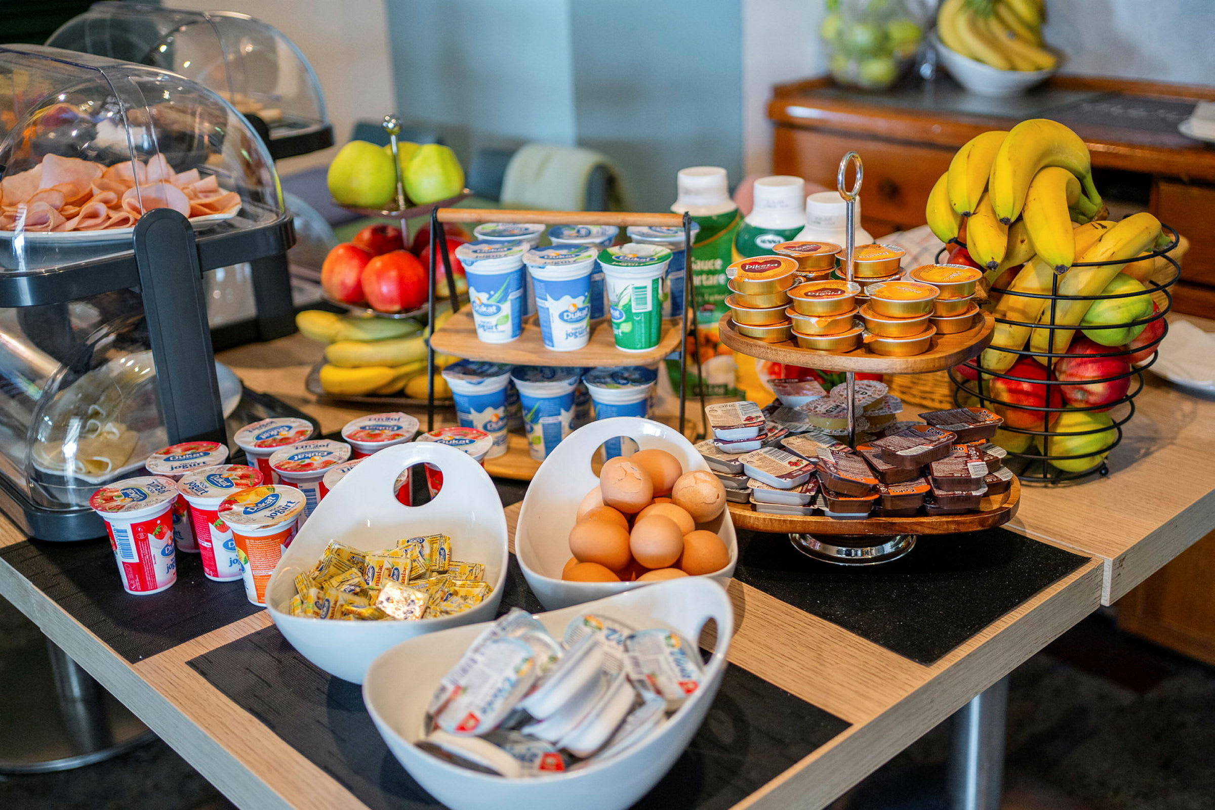 Bildet viser et frokostbord med forskjellige matvarer som egg, yoghurt, frukt, pålegg og smørpakker. (AI-generert tekst)