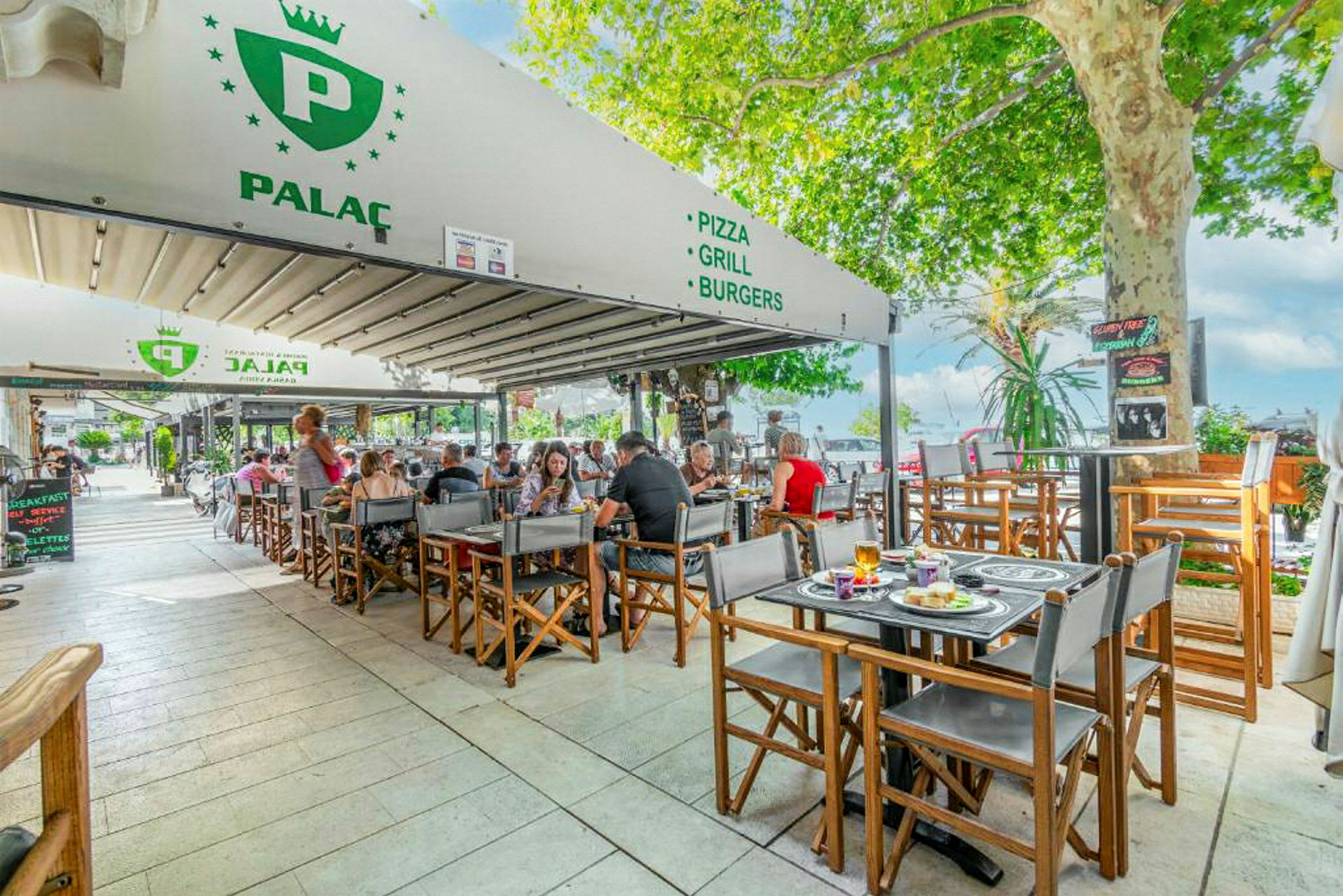 Uteservering ved restaurant med gjester ved bord, omgitt av grøntområder. (AI-generert tekst)