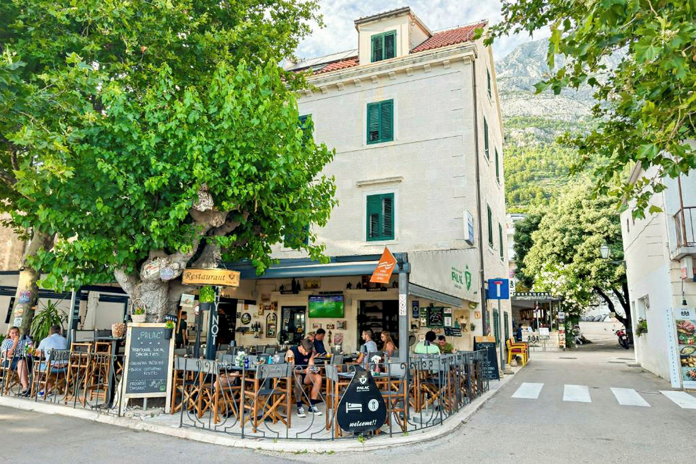 En utendørs kafé ved en bygning med hvite vegger og grønne skodder, omgitt av grøntområder og trær. (AI-generert tekst)