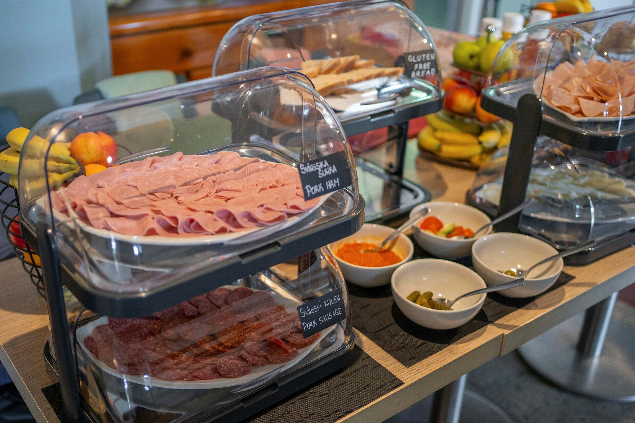 Buffetbord med skiver av skinke og pølse, et utvalg av dressinger og grønnsaker samt frukt i bakgrunnen. (AI-generert tekst)