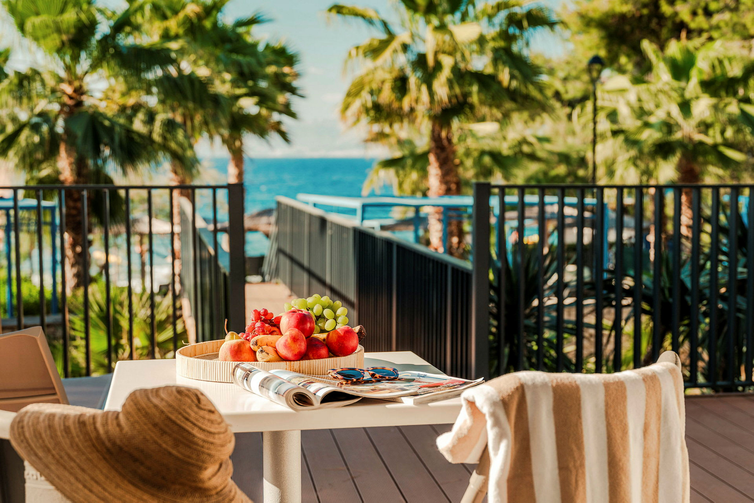 Et dekket bord med fruktfat, en avis og et par solbriller, med utsikt til en terrasse med palmer og havet i bakgrunnen. (AI-generert tekst)