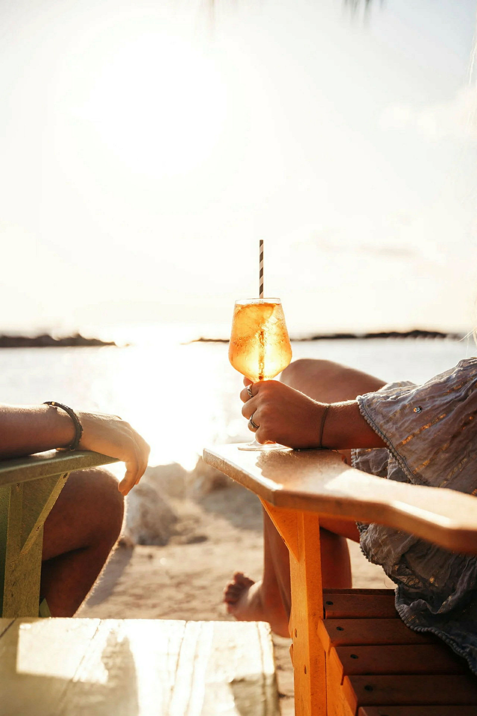 To personer sitter i stoler på en strand med utsikt over havet og solnedgangen. I forgrunnen holdes et glass med en drink. (AI-generert tekst)