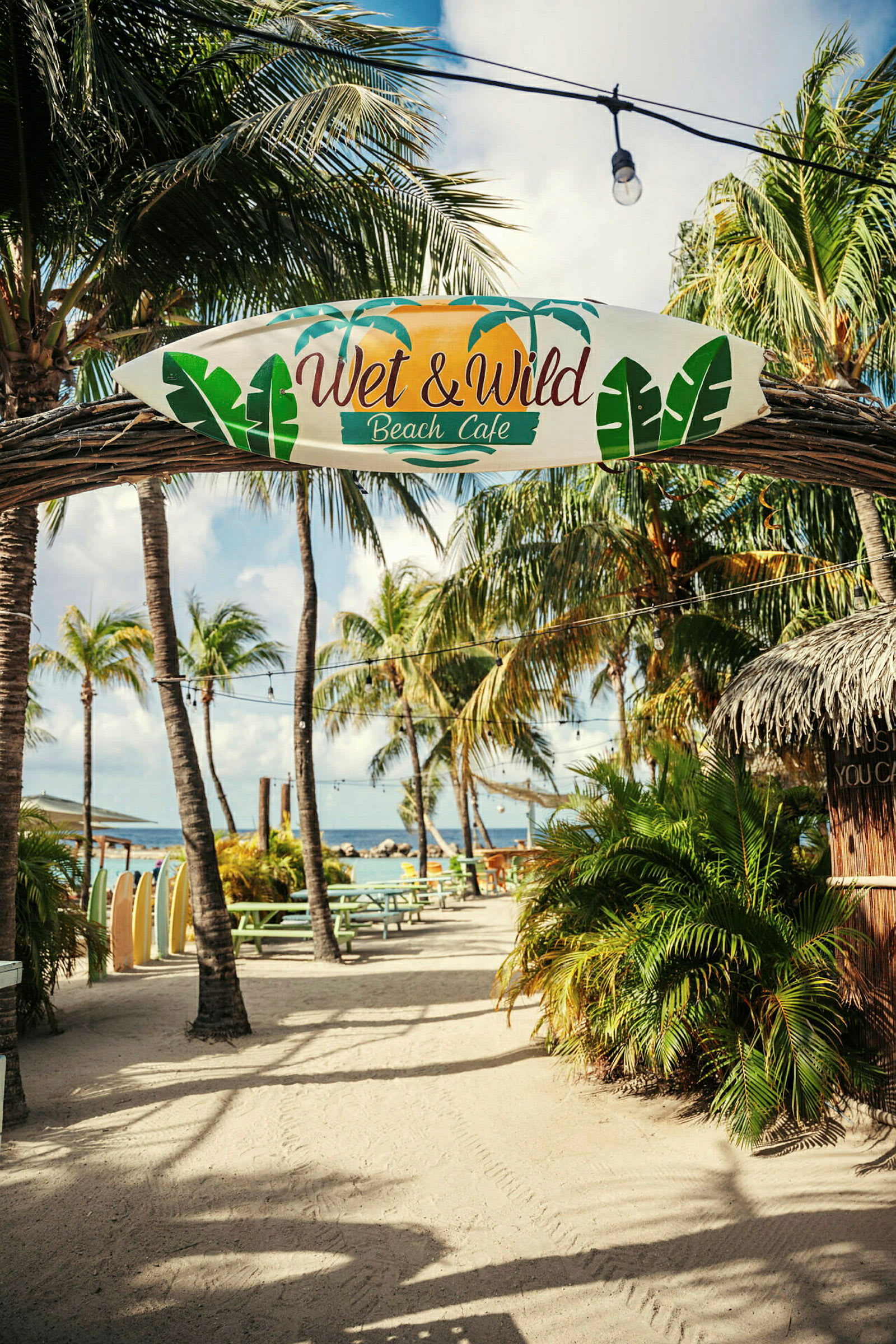 Skilt med teksten "Wet & Wild Beach Café" henger på en bue i et utendørsmiljø med palmer, bord og hav i bakgrunnen. (AI-generert tekst)