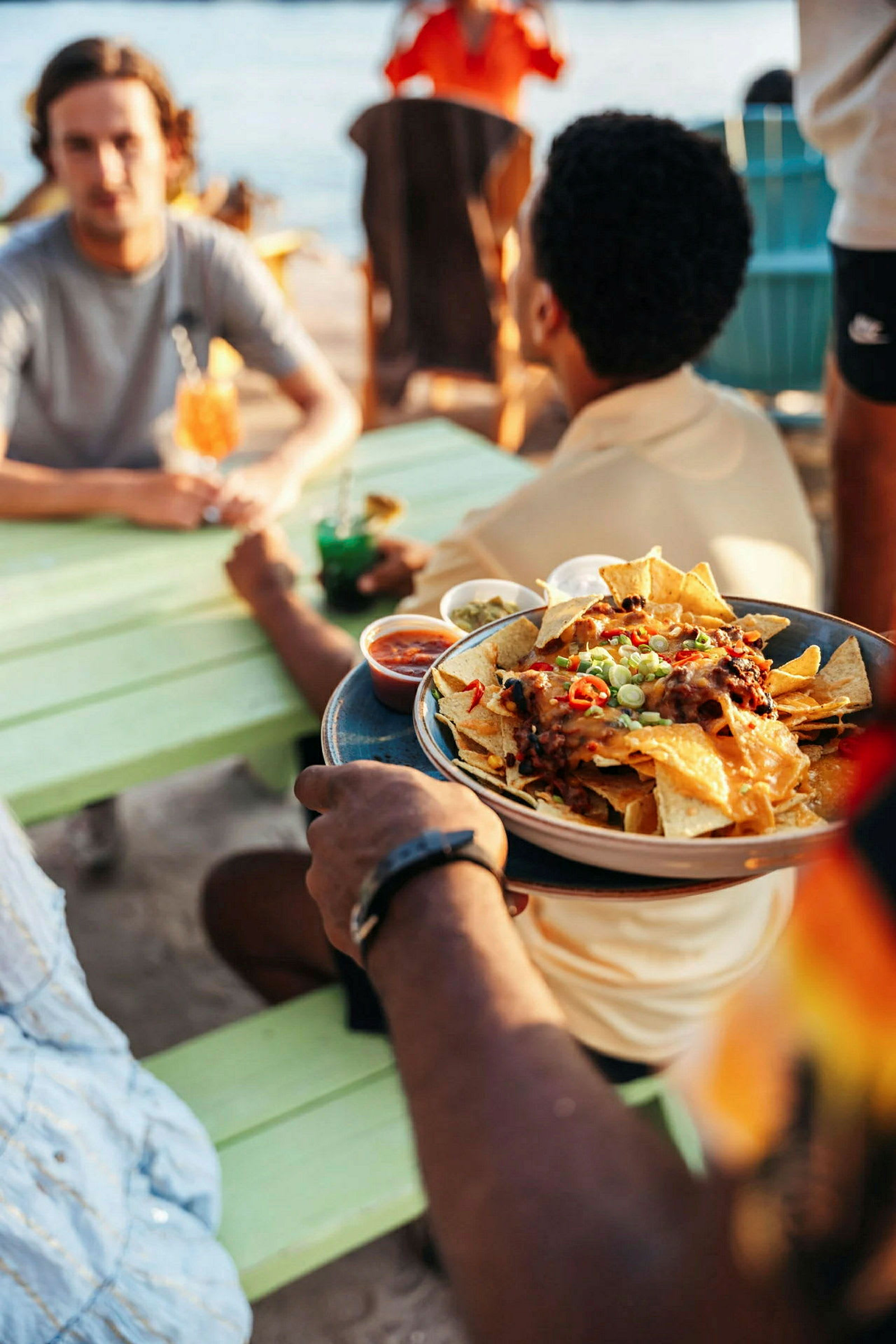 En person holder en tallerken med nachos, ulike dippesauser vises rundt tallerkenen. Scenen er utendørs ved et bord nær vann. (AI-generert tekst)