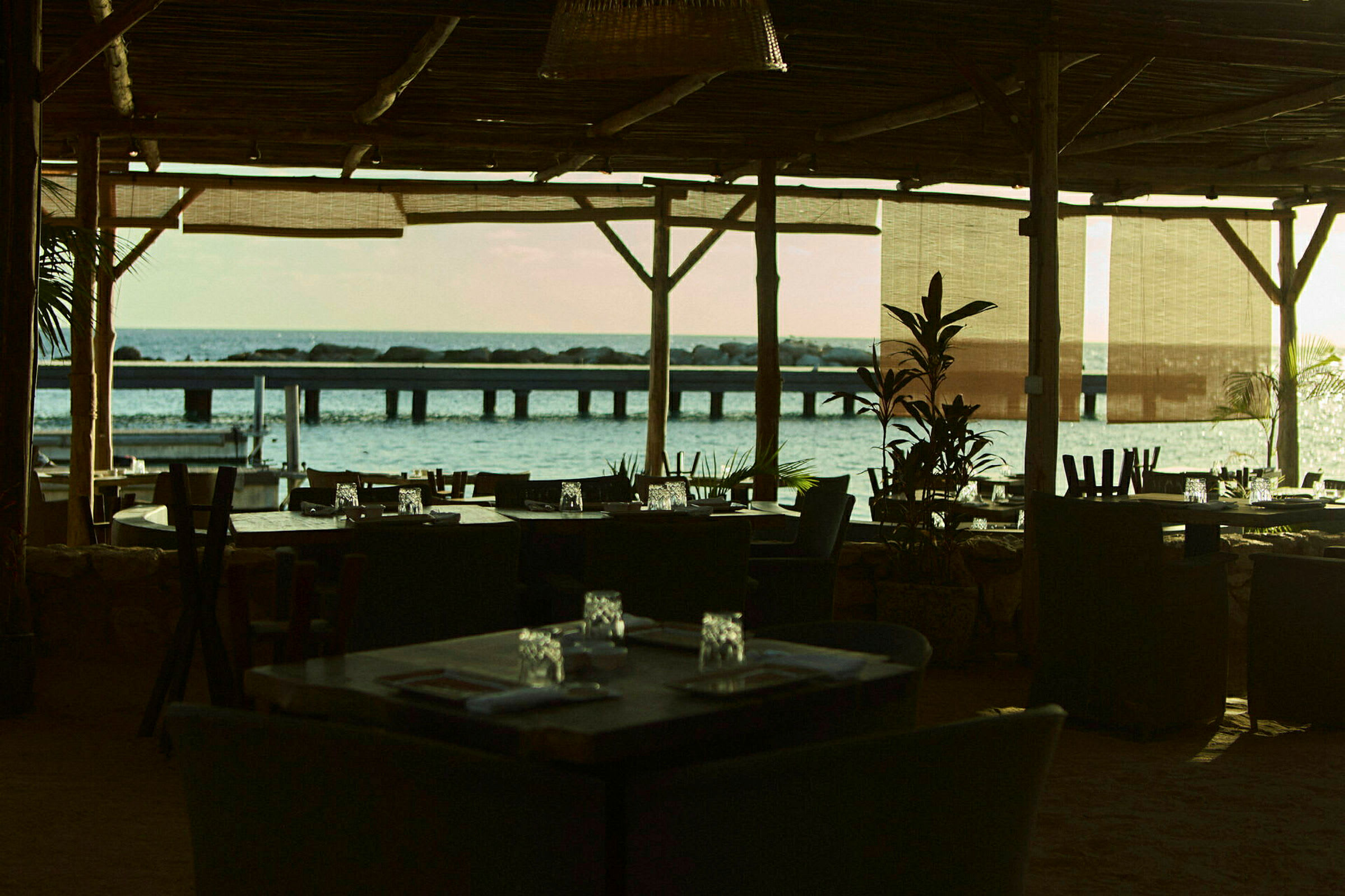 En restaurant med tak og møbler med utsikt over havet og en brygge i bakgrunnen. (AI-generert tekst)