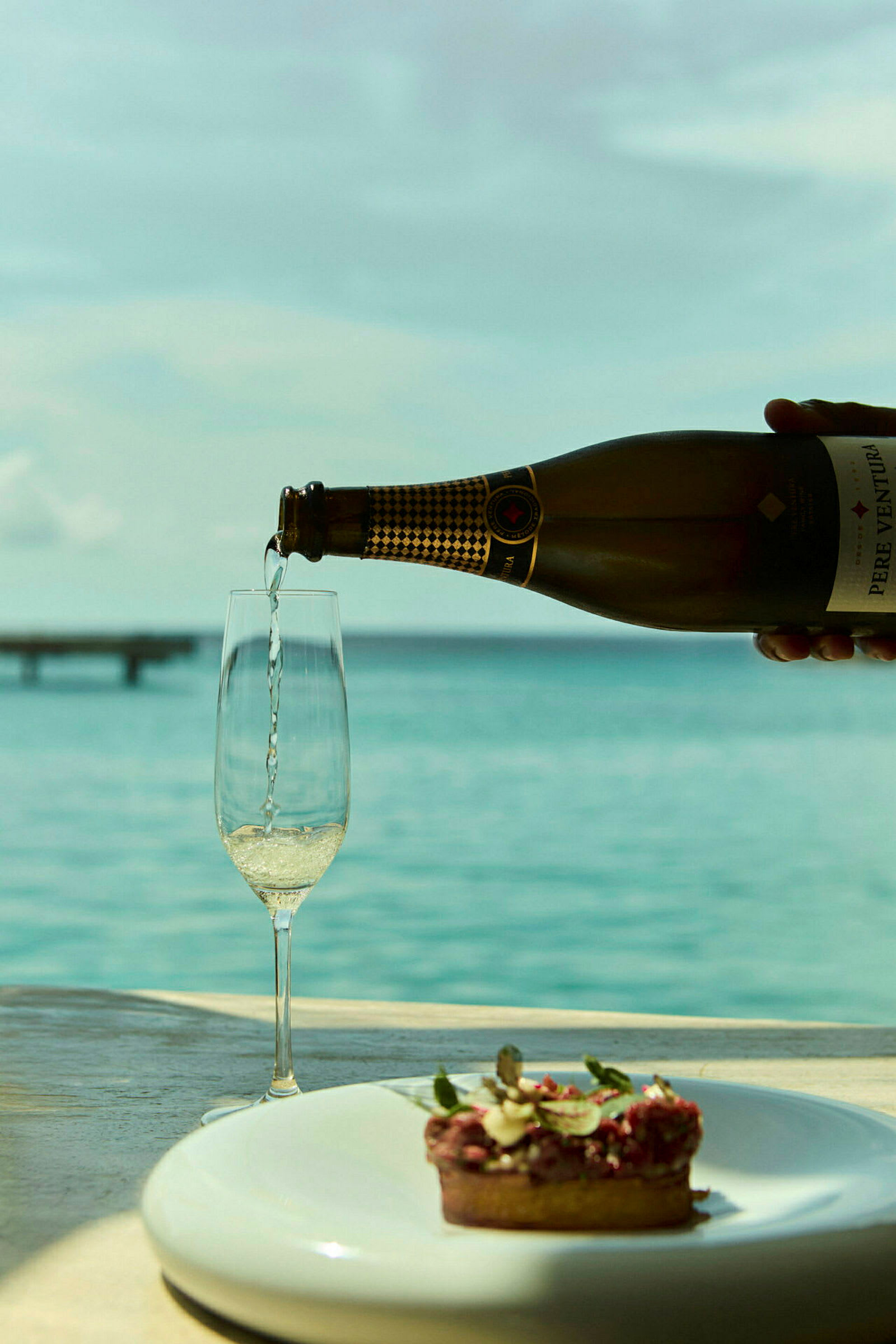 Et glass fylles med champagne ved siden av en tallerken med en liten dessert, med havutsikt i bakgrunnen. (AI-generert tekst)