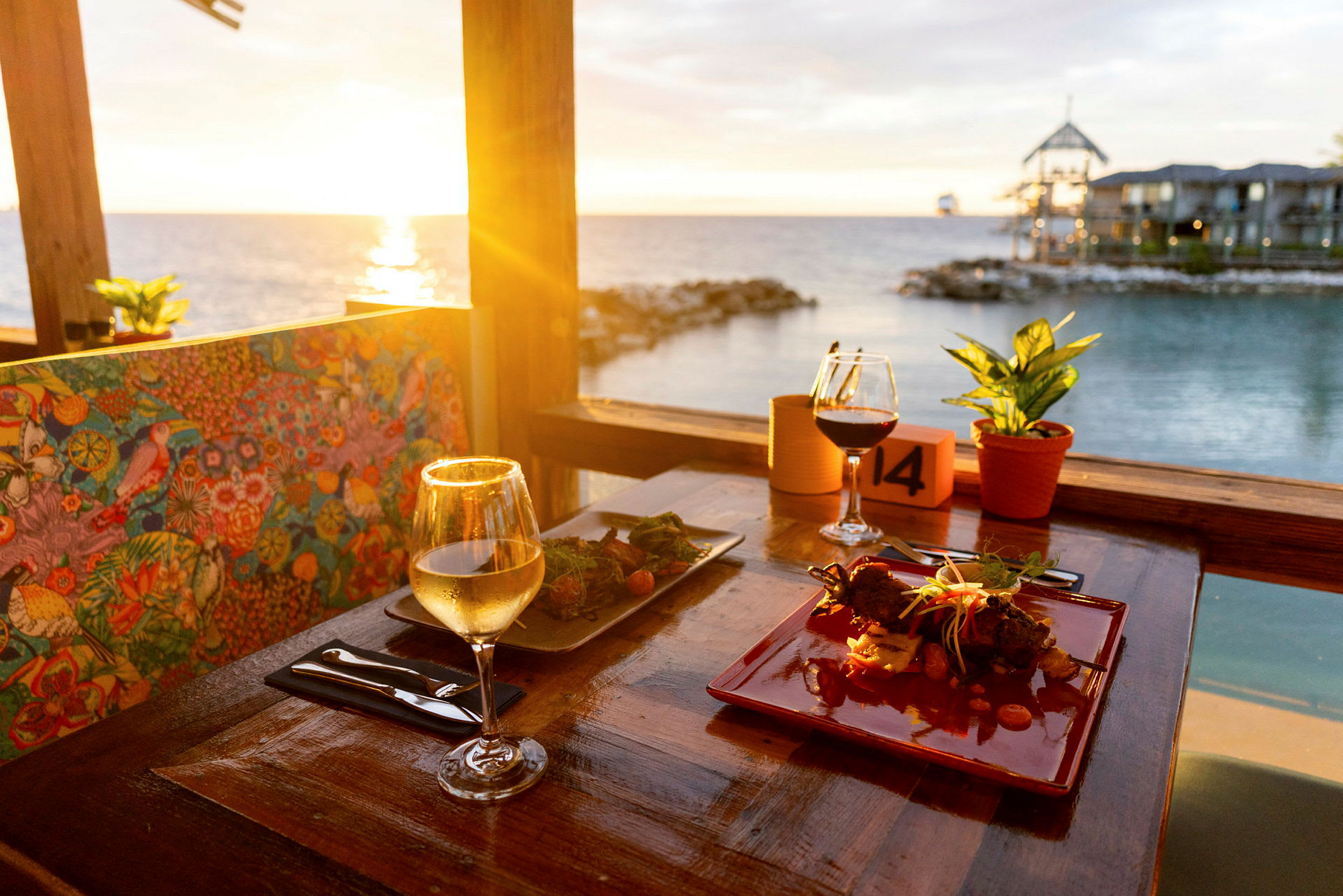 En restaurantstolrygg med fargerikt mønster, et veldesignet bord med vinglass og mat, utsikt over havet ved solnedgang. (AI-generert tekst)