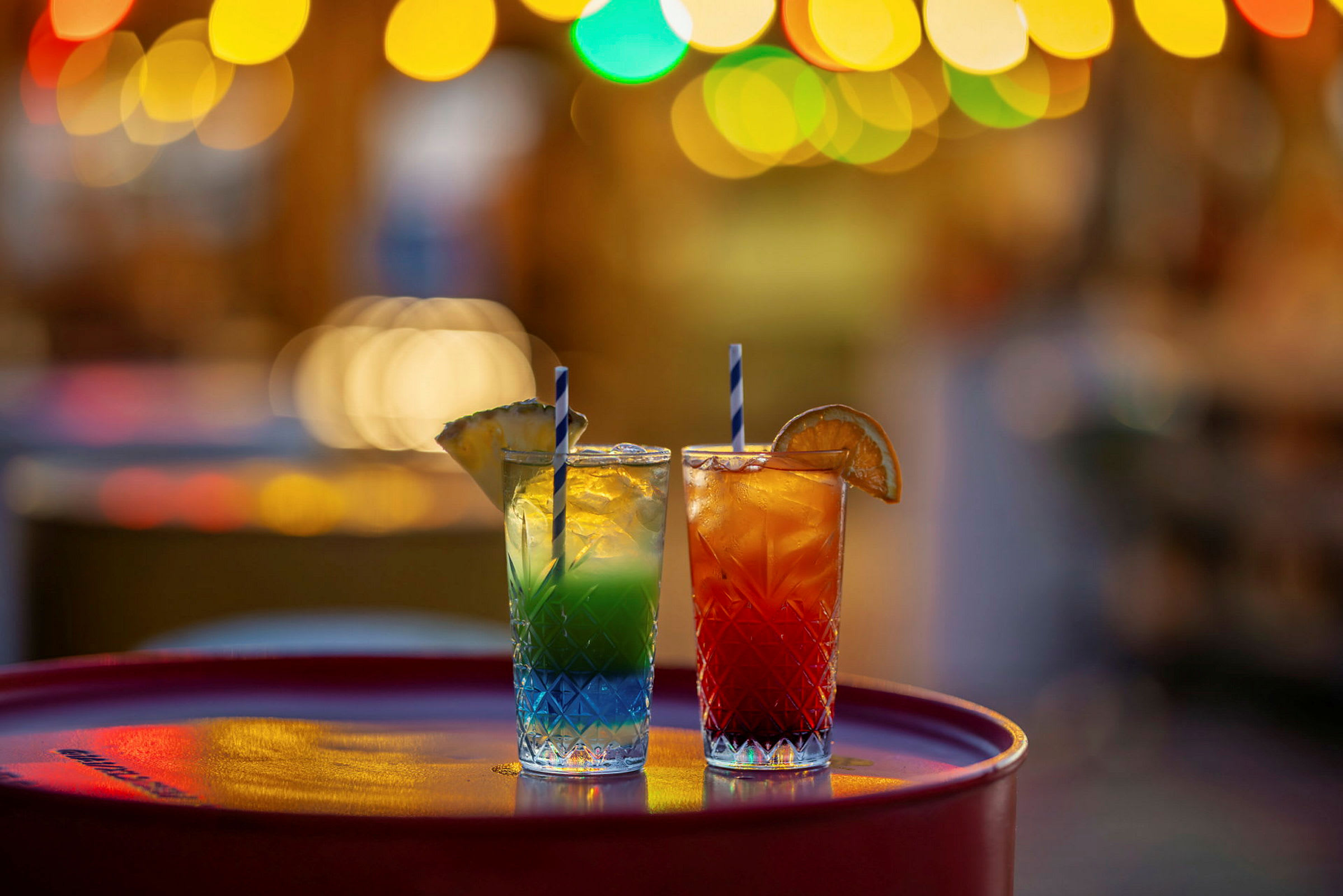 To glass med fargerike cocktails, dekorert med frukt, står på et bord med uskarpe lys i bakgrunnen. (AI-generert tekst)