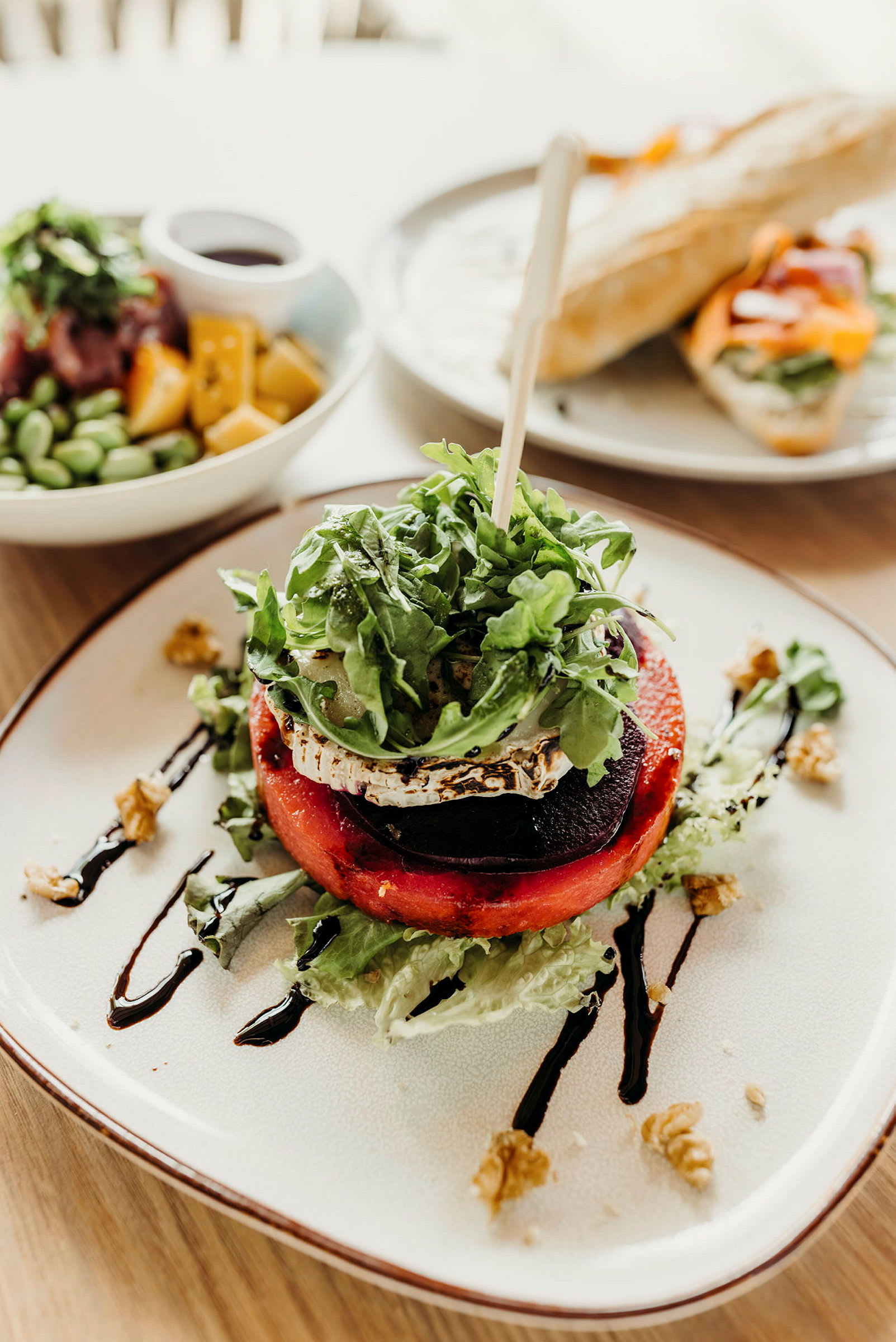 En tallerken med en salat som inneholder grillet grønnsak, ost og ruccola, dekorert med balsamico og nøtter. I bakgrunnen sees en brødrett og en skål med fargerike ingredienser. (AI-generert tekst)
