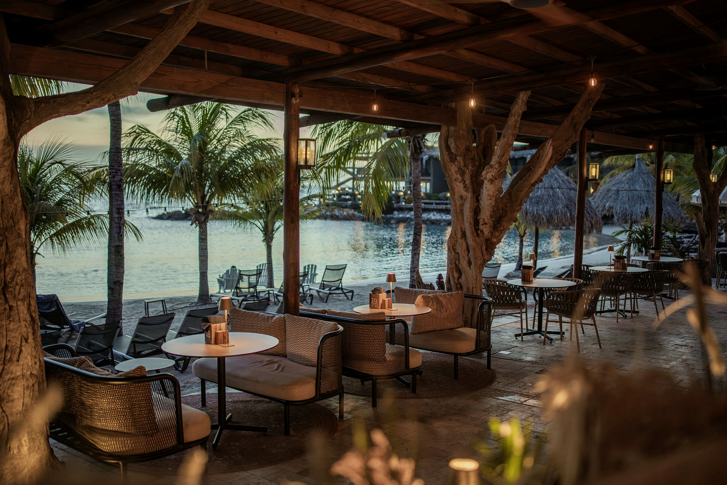 En utendørs lounge under et tretak med sofaer og bord ved havet, omgitt av palmetrær. (AI-generert tekst)