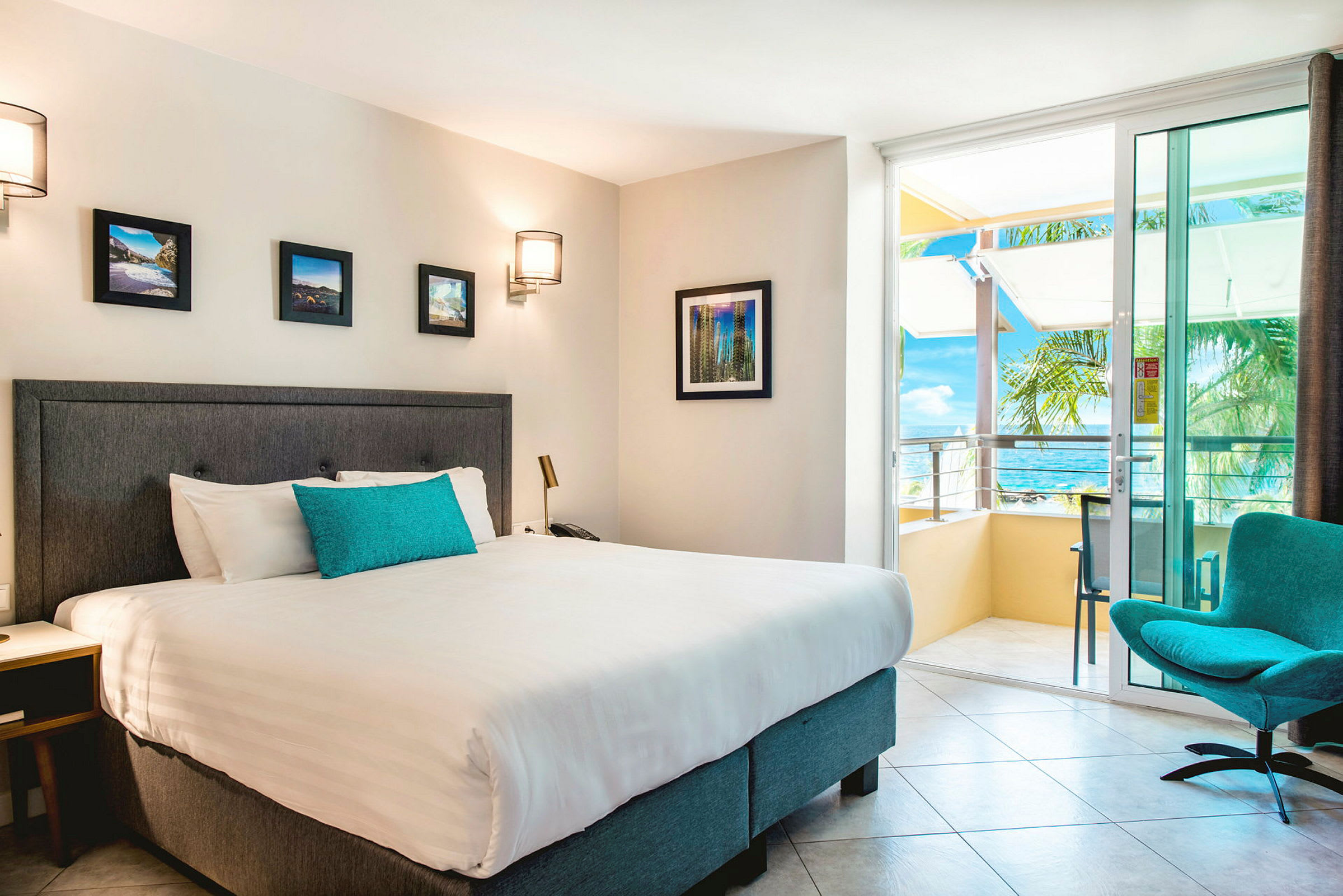 Hotellrom med en dobbeltseng, veggkunst og en stol ved siden av en balkong med havutsikt. (AI-generert tekst)