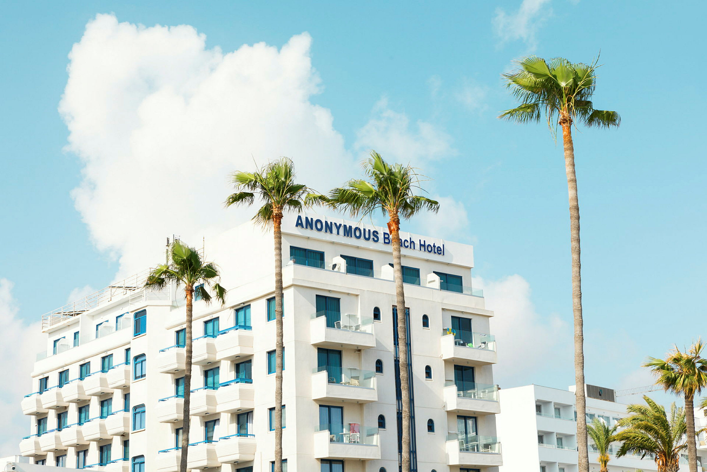 Et hotell med navnet Anonymous Beach Hotel, hvit fasade og blå vinduer, omgitt av palmer. (AI-generert tekst)