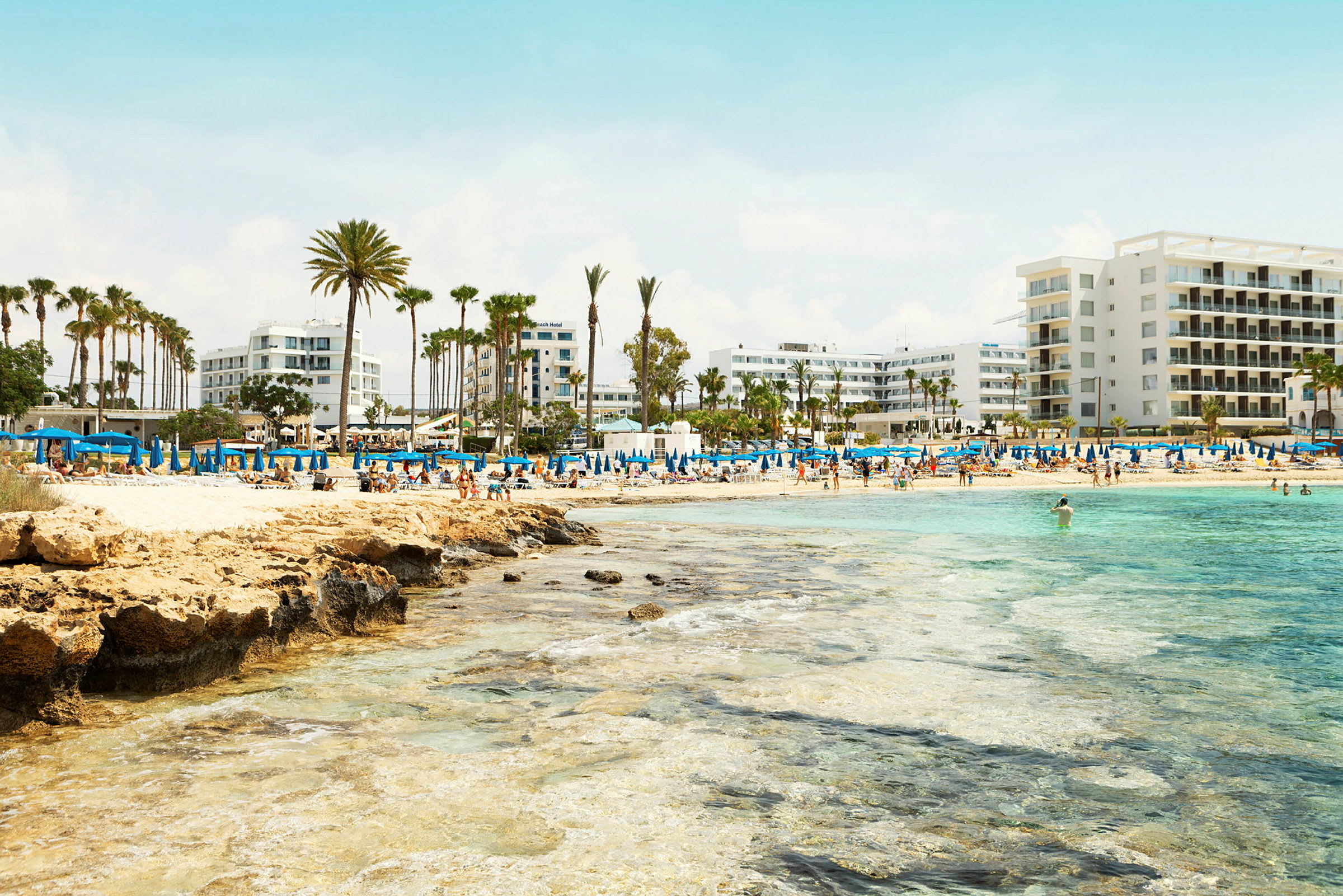 Strand med blå parasoller, palmer og hotellbygninger i bakgrunnen. (AI-generert tekst)