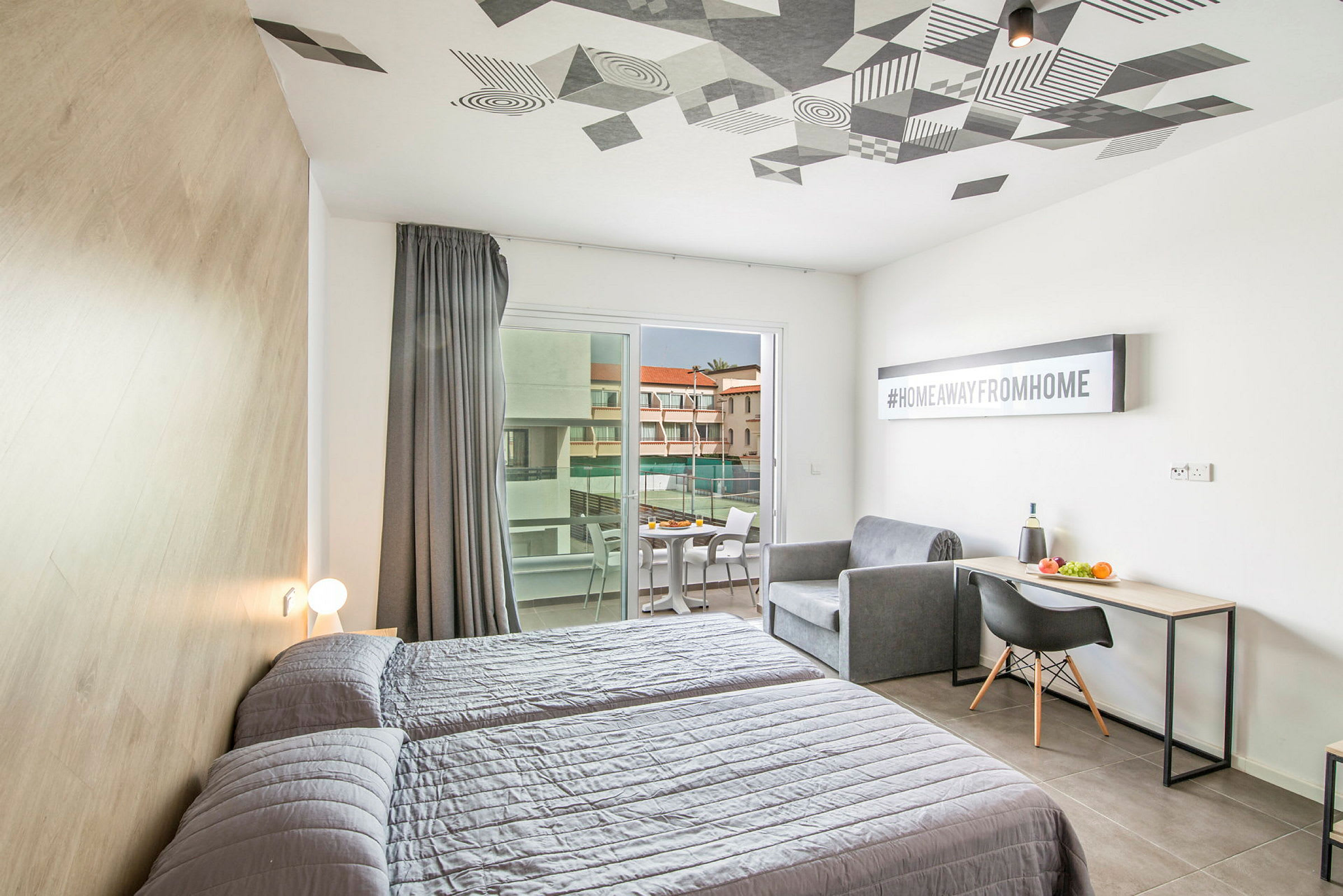 Et hotellrom med to senger, en sofa, et skrivebord og en balkong med utsikt mot en bygning. (AI-generert tekst)