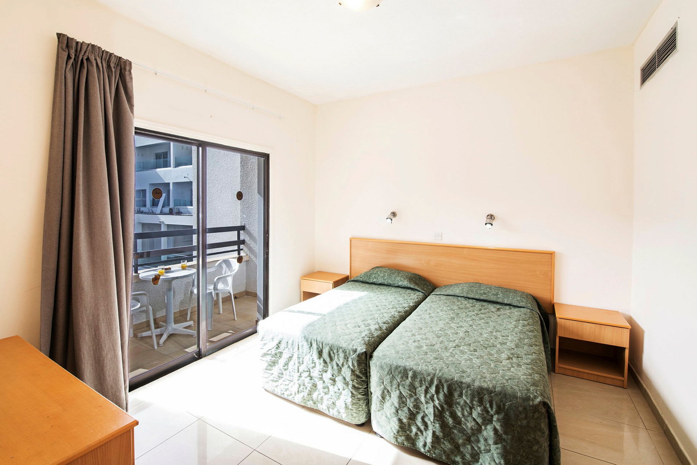 To enkeltsenger i et lyst hotellrom med balkong og sittemøbler utenfor. (AI-generert tekst)
