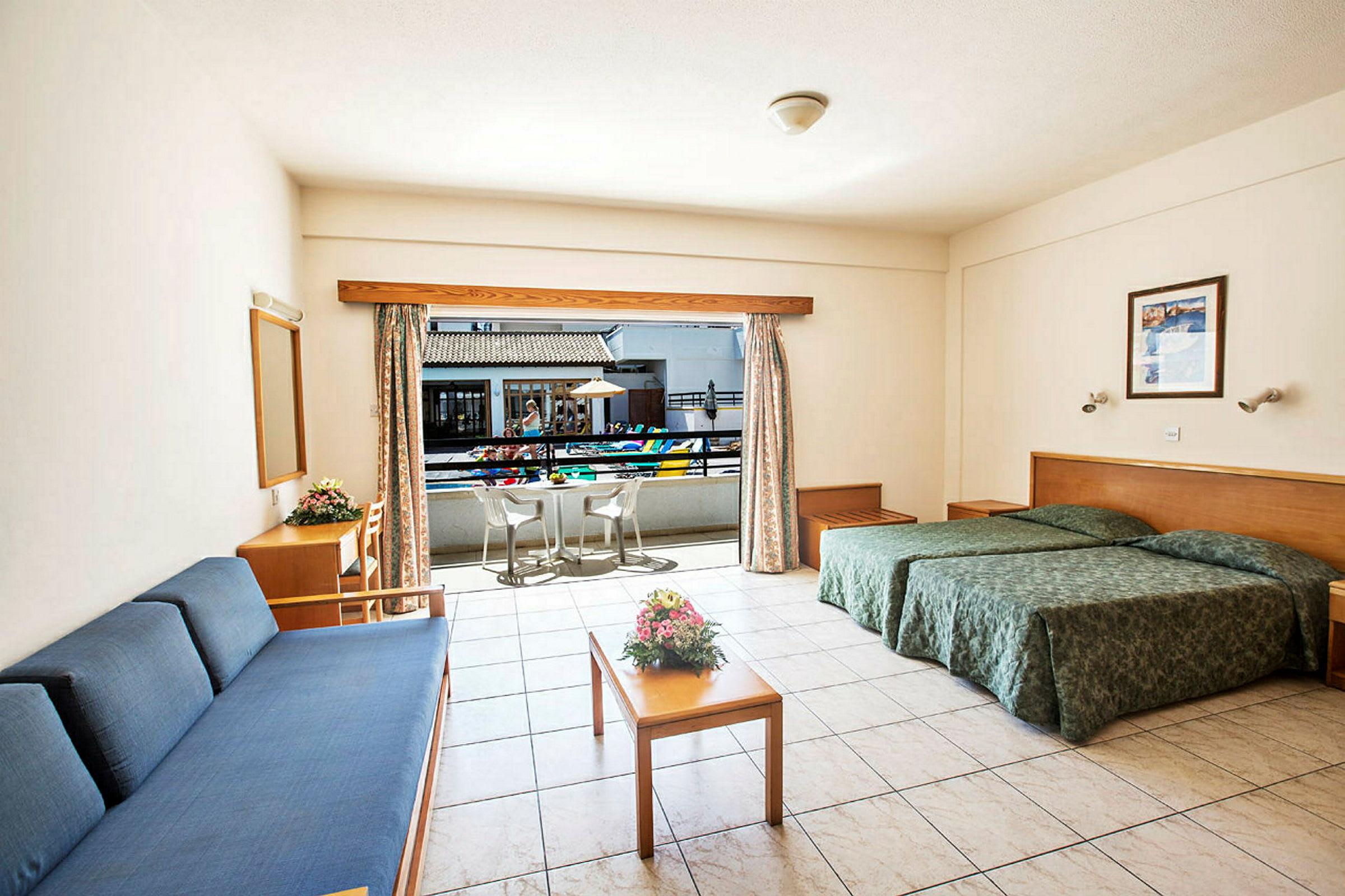 Hotellrom med to enkeltsenger, sofa, bord med blomsterbukett og utgang til balkong med stoler. (AI-generert tekst)