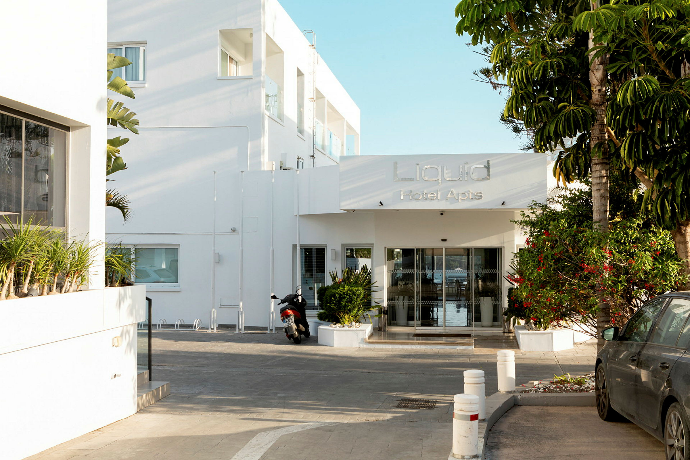 Inngang til Liquid Hotel Apartments med en motorsykkel og bil foran. (AI-generert tekst)