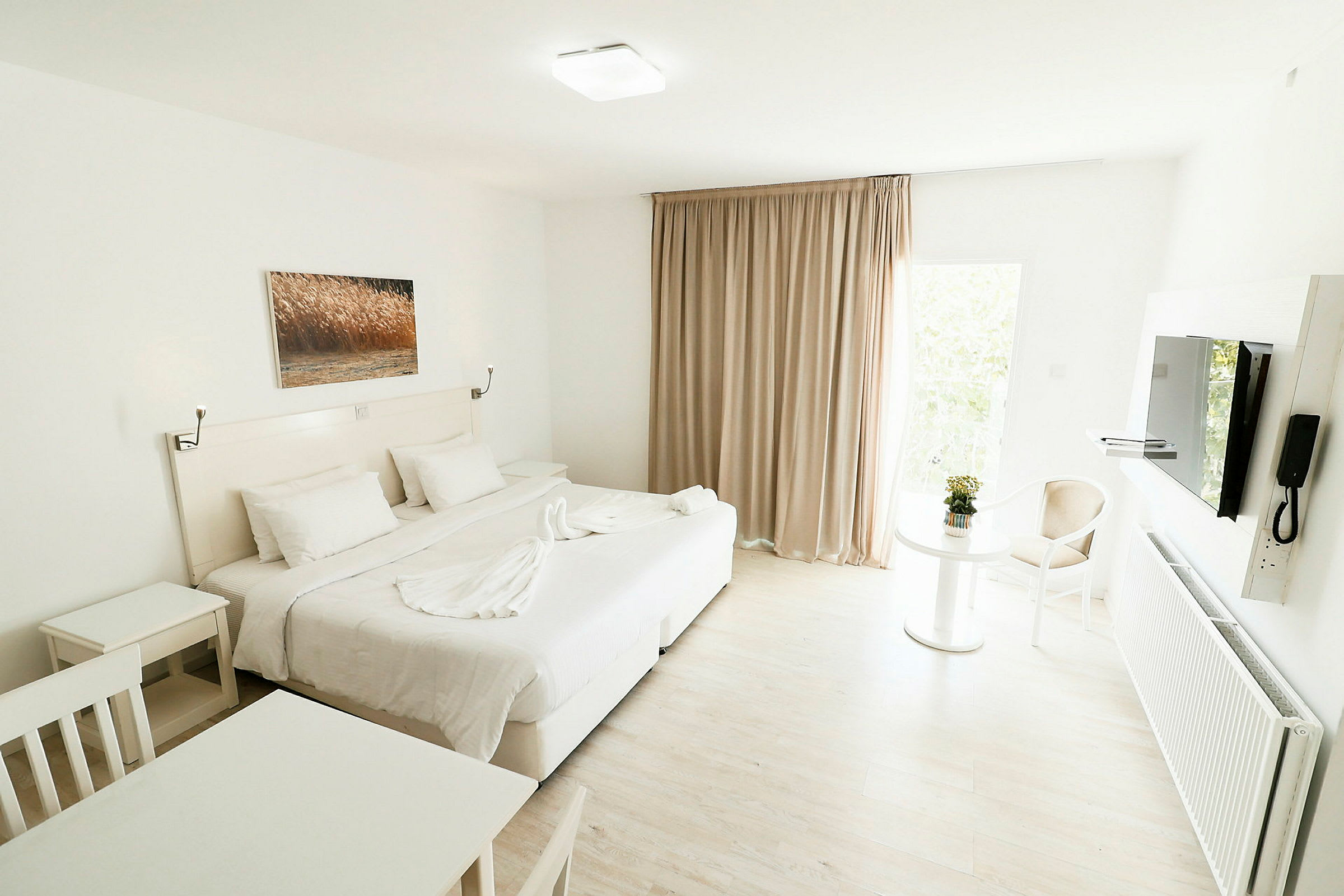 Et lyst og moderne hotellrom med en stor seng, hvite møbler og et bilde på veggen. Et bord med en blomst og en stol er synlig ved et vindu med beige gardiner. (AI-generert tekst)