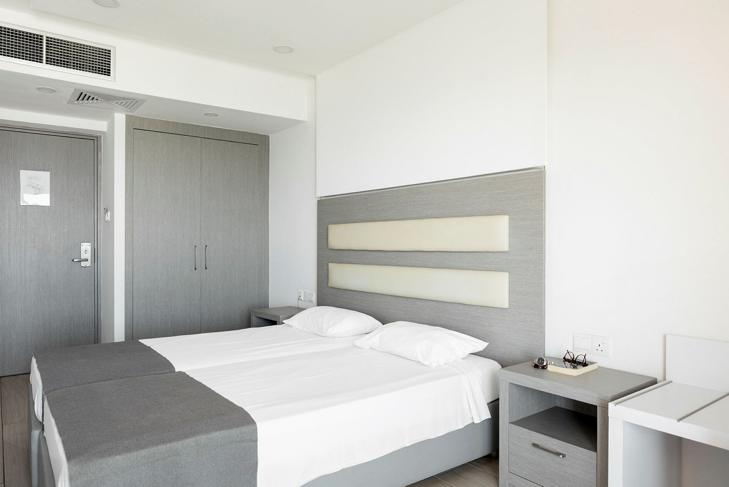 Bildet viser et hotellrom med to enkeltsenger, grå innredning og hvite vegger. (AI-generert tekst)