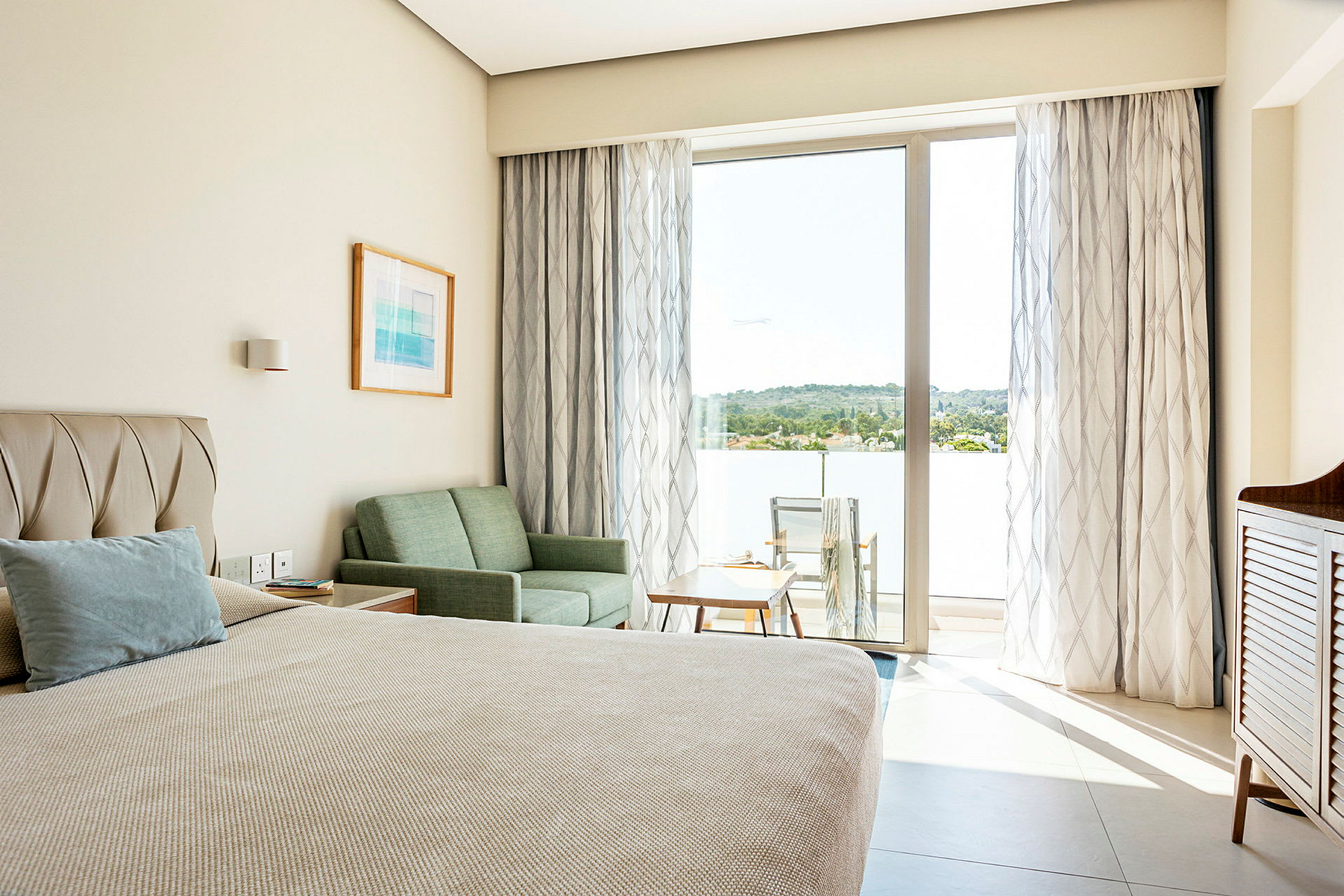 Et hotellrom med en seng, sofa og bord, samt en balkong med stoler og utsikt over grøntområder. (AI-generert tekst)