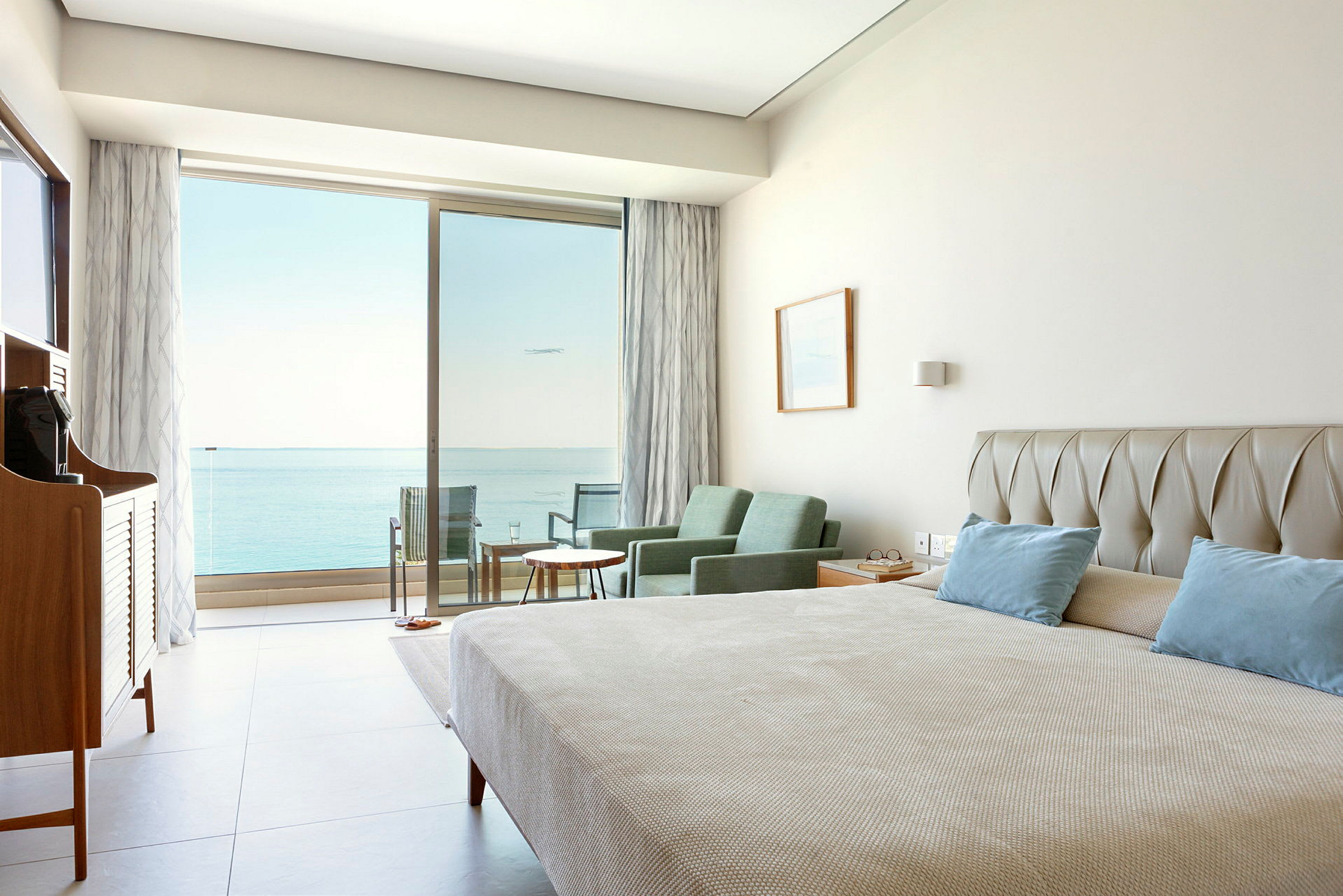 Hotellrom med seng, sittegruppe og utsikt mot havet gjennom glassdører. (AI-generert tekst)