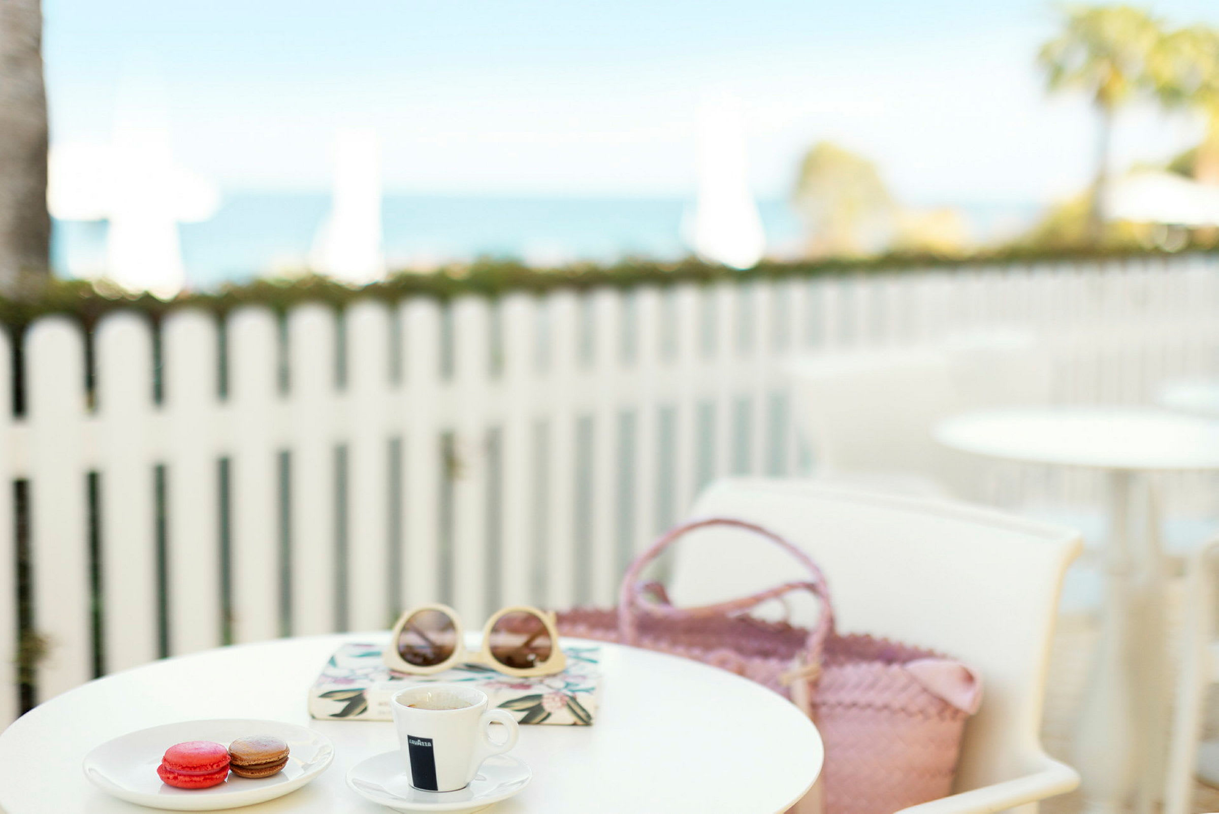 En kopp kaffe, macarons, solbriller og en veske på et kafébord med et hvitt tregjerde og havet i bakgrunnen. (AI-generert tekst)