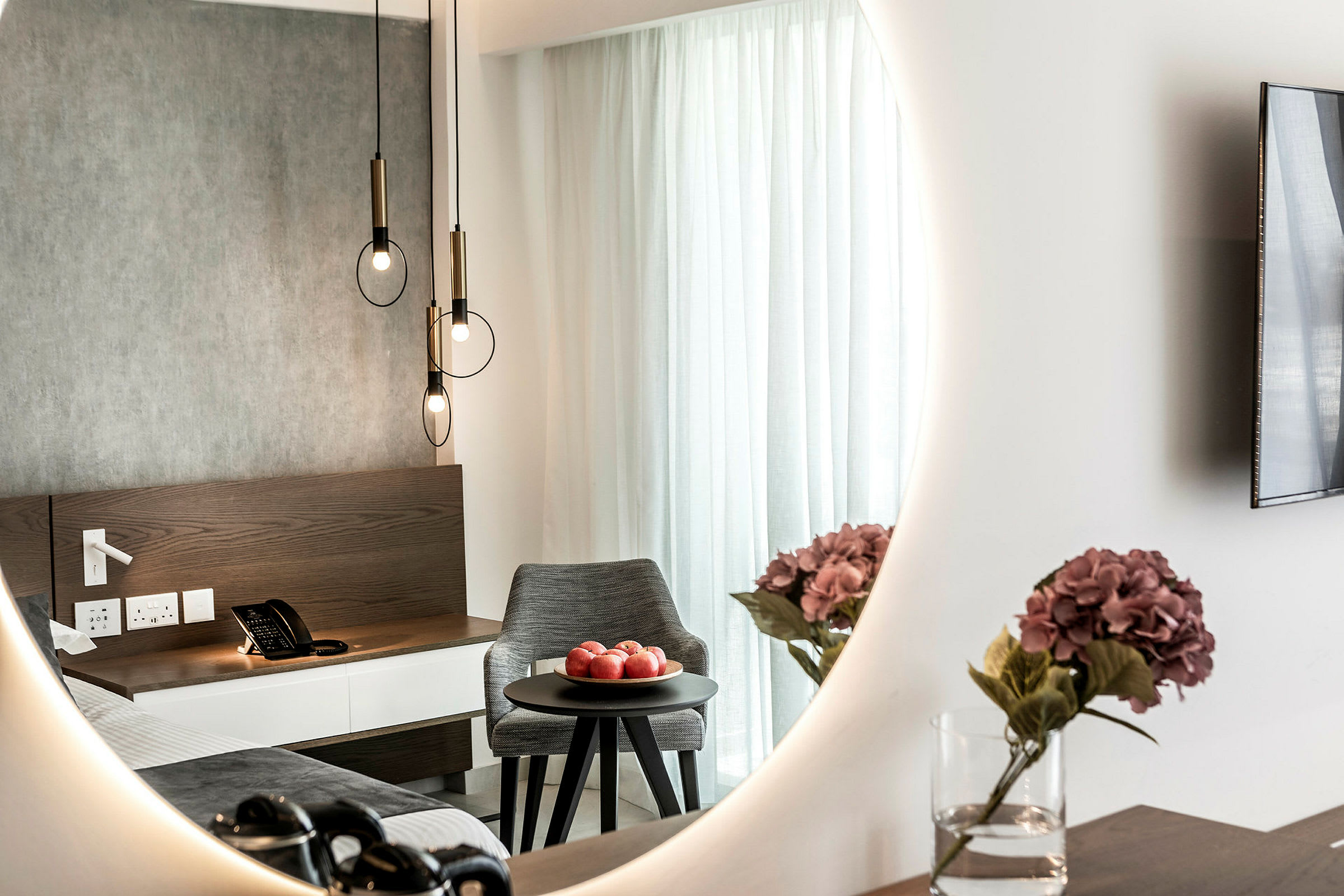Interiør av hotellrom med et speil som reflekterer en seng, et nattbord med en telefon, en skål med røde epler på et rundt bord og en vase med lilla blomster. (AI-generert tekst)
