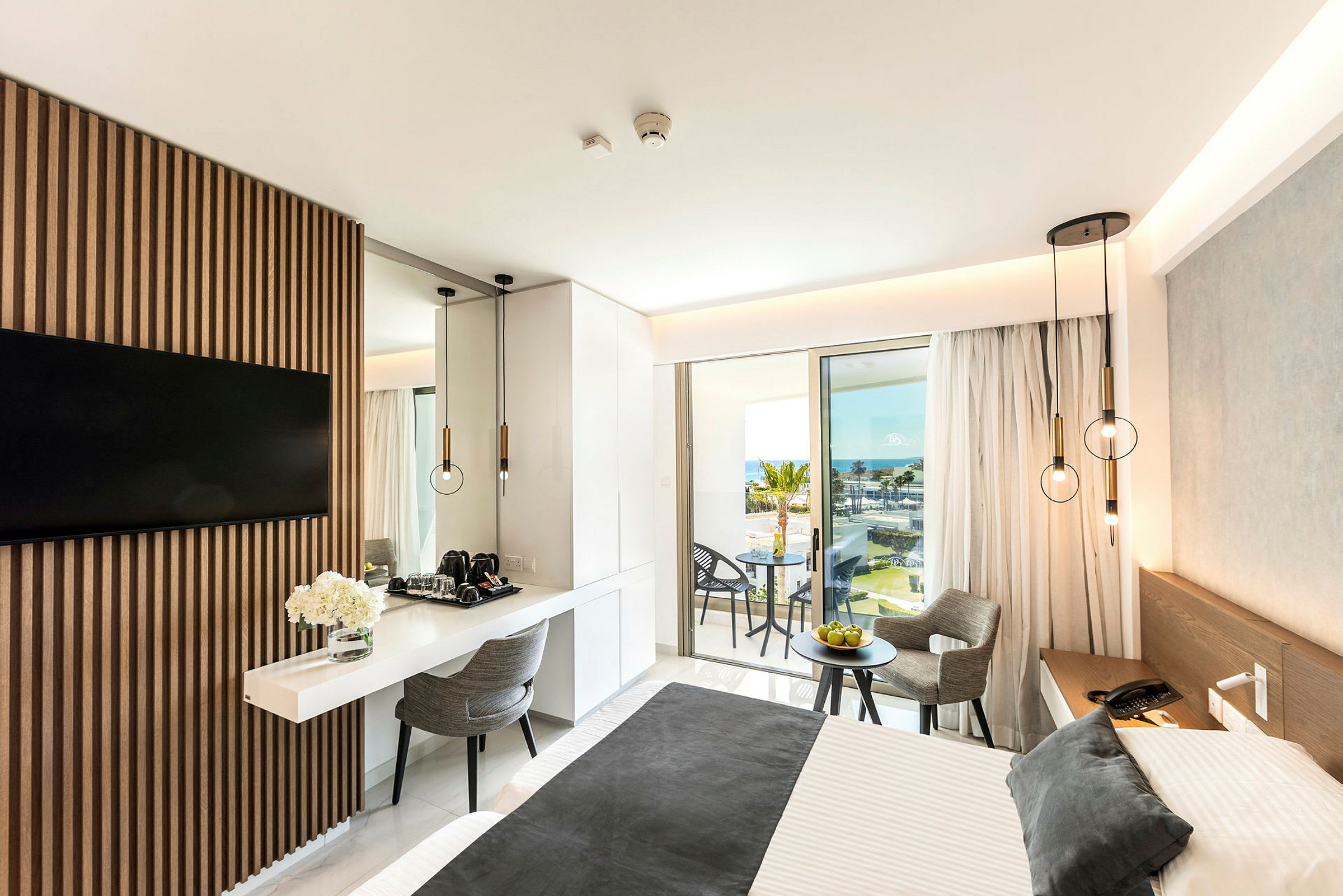 Moderne hotellrom med en dobbeltseng, bord og stoler samt utsikt over havet fra en balkong. (AI-generert tekst)