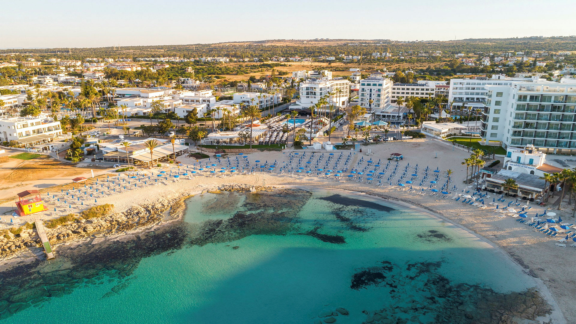 Strand med parasoller og stoler, omgitt av turkis vann, bygninger og grøntområder. (AI-generert tekst)