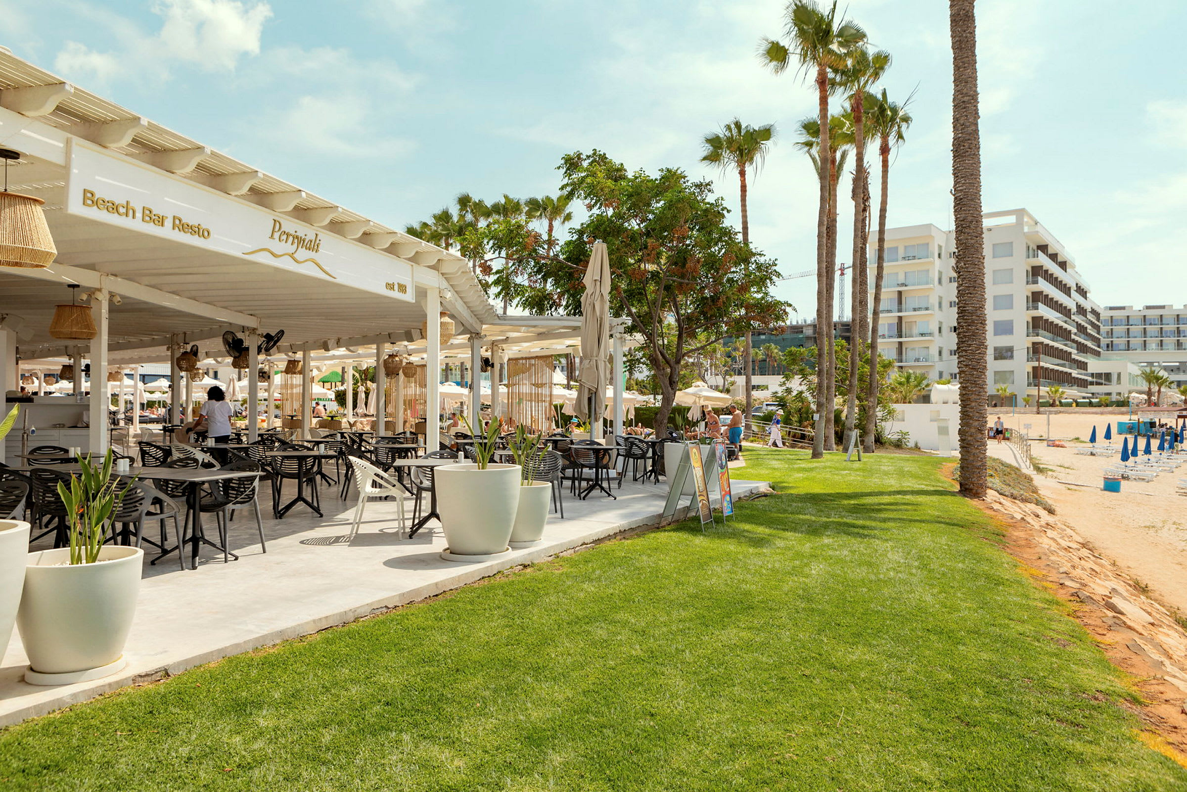 Bilde av en strandbar med sitteplasser og parasoller samt hotellbygninger i bakgrunnen. (AI-generert tekst)