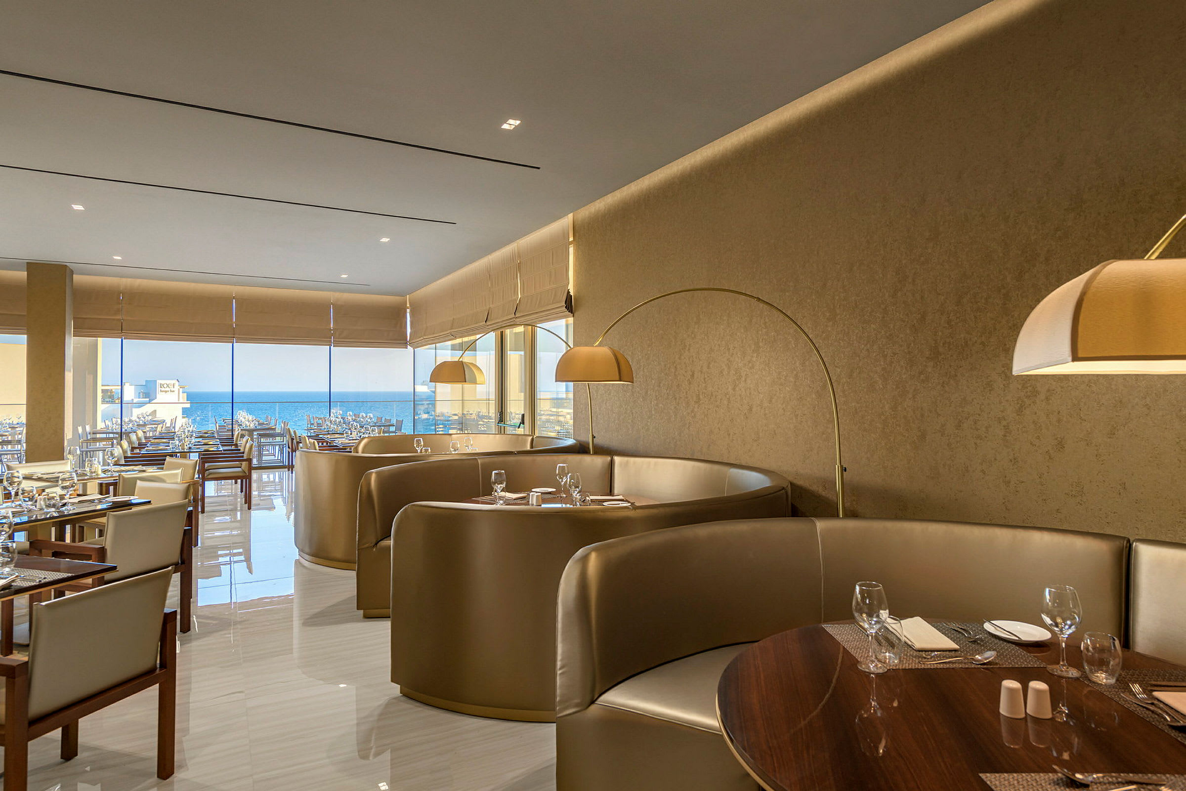 En elegant restaurant med sittegrupper og bord dekket med glass og bestikk samt utsikt over havet. (AI-generert tekst)
