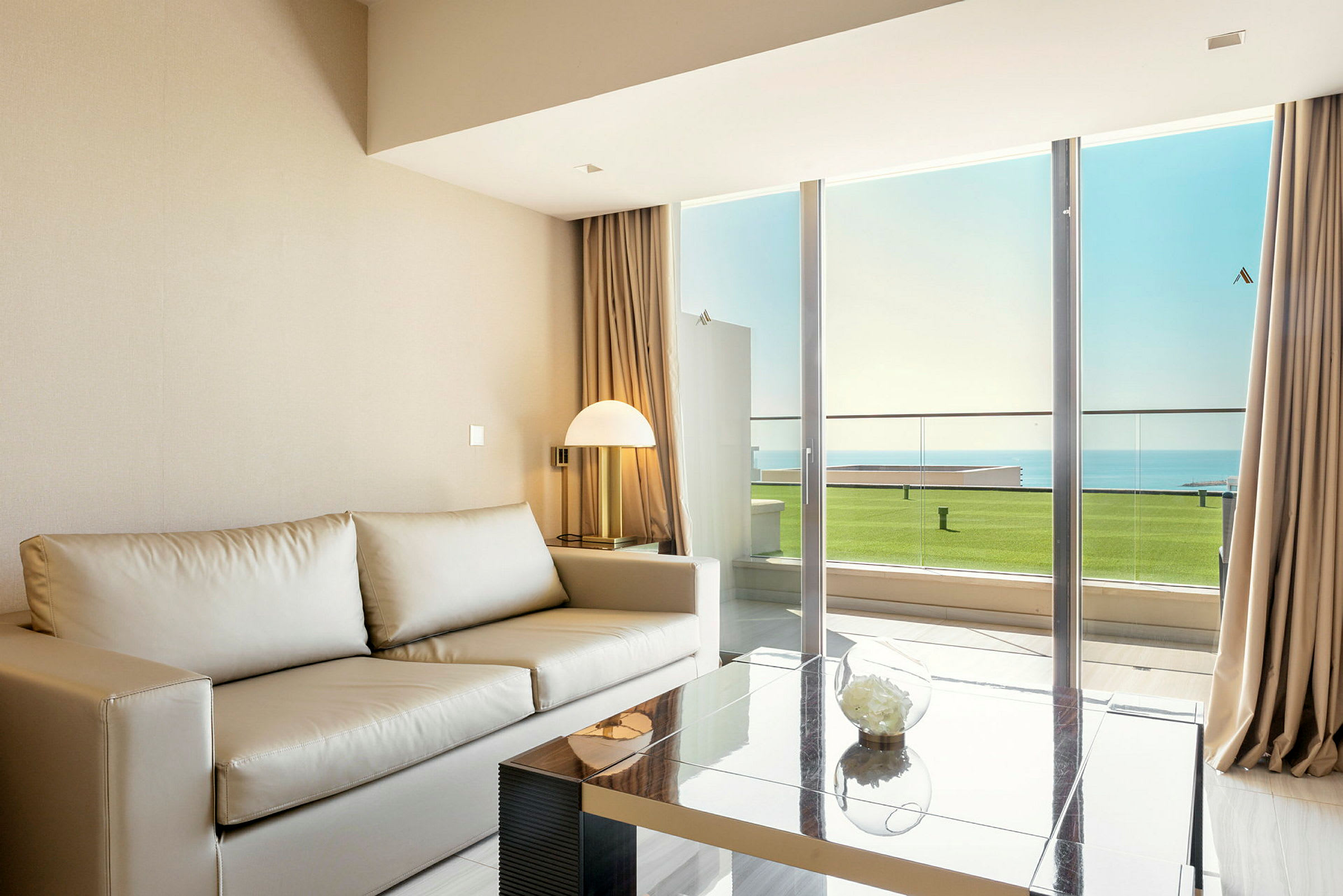 Bilde av en moderne stue med en beige sofa, glassbord og en stor vindusdør som vender mot en balkong med utsikt mot havet. (AI-generert tekst)