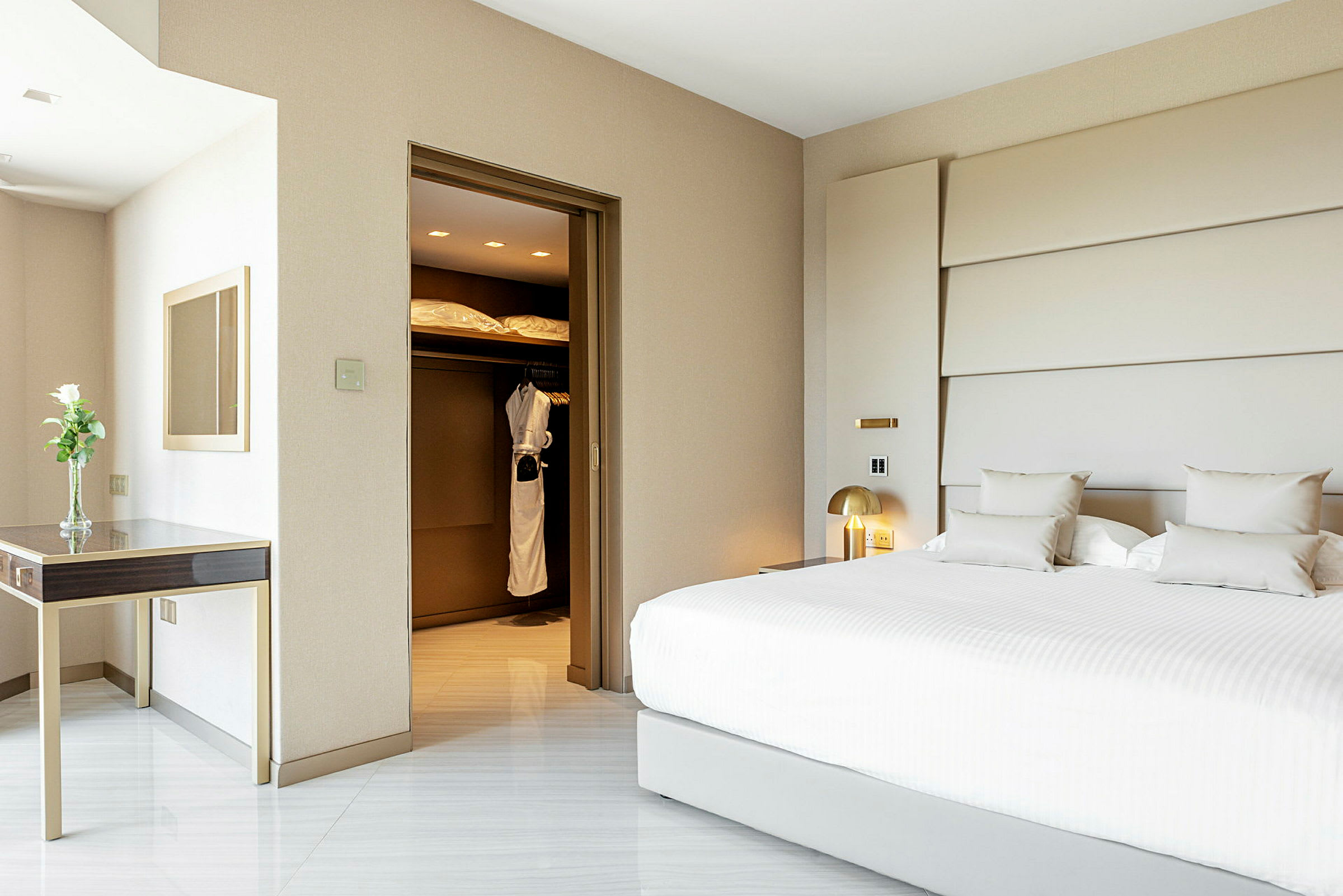 Hotellrom med en stor hvit seng, et skrivebord med en vase og et skap som inneholder badekåper. (AI-generert tekst)