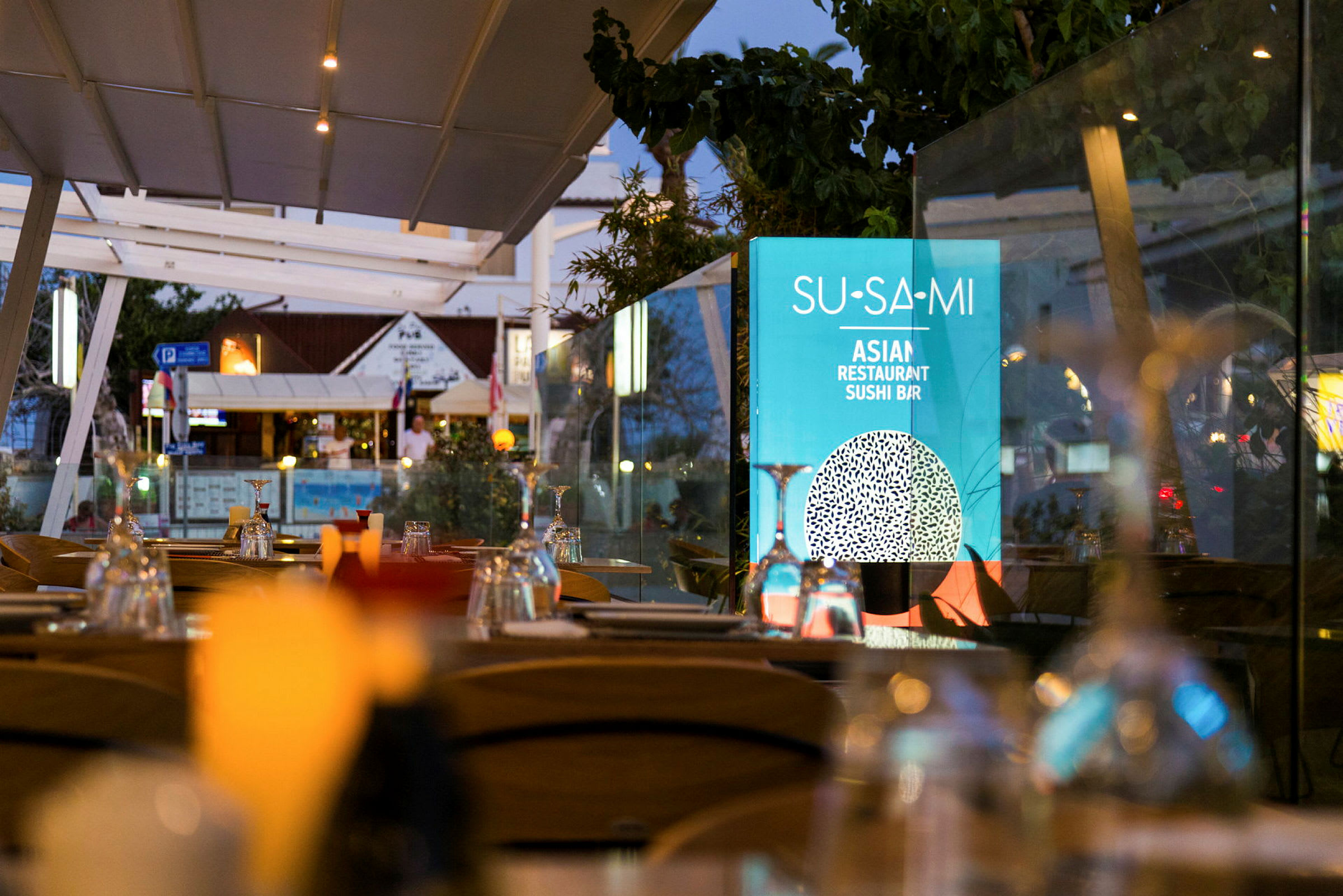 Uteservering med dekkede bord og en skilt med teksten 'Su-Sa-Mi Asian Restaurant Sushi Bar'. (AI-generert tekst)