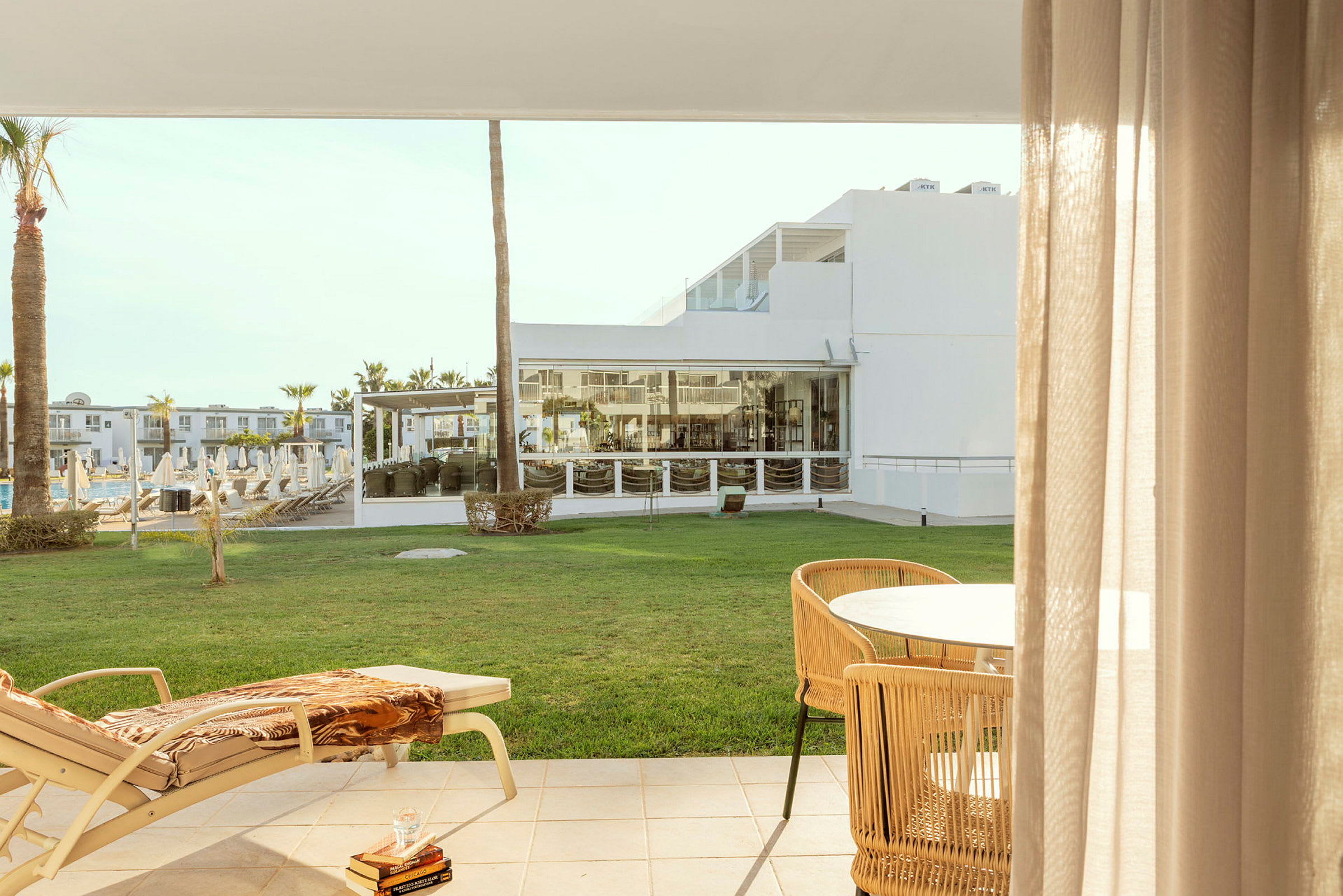 Utsikt fra en terrasse med en solseng og bord, med en grønn plen og hotellbygninger i bakgrunnen. (AI-generert tekst)
