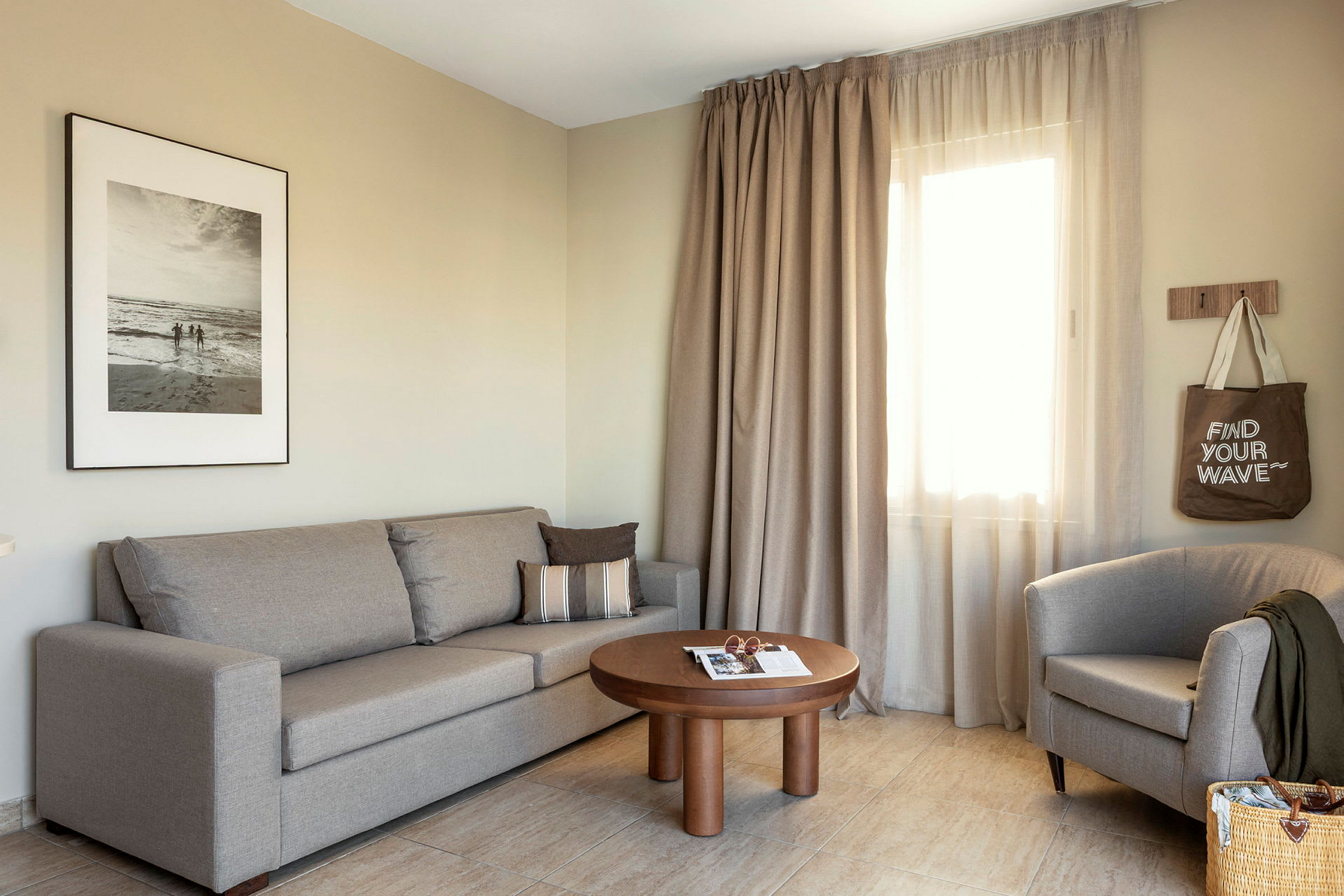 Et stue med en beige sofa, en lenestol, et trebord og gardiner ved et vindu. (AI-generert tekst)