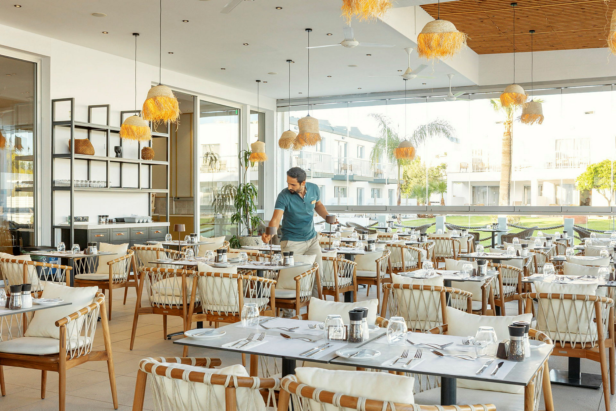 Restaurant med bord og stoler samt store vinduer som vender ut mot et grønt uteområde. (AI-generert tekst)
