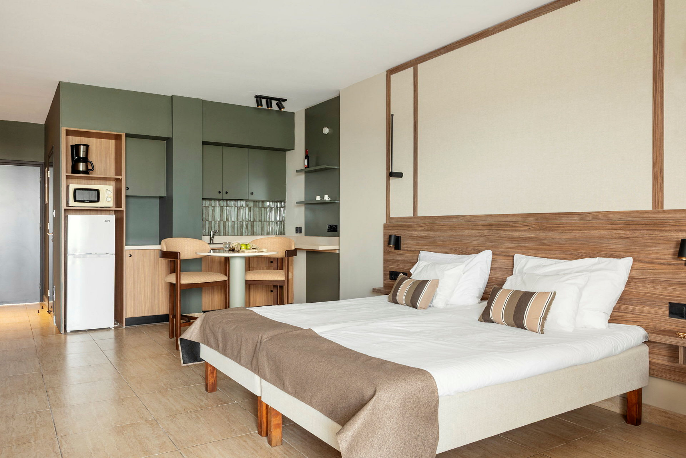Innendørs bilde av et lyst og moderne hotellrom med en seng, bord, stoler og kjøkkenområde. (AI-generert tekst)