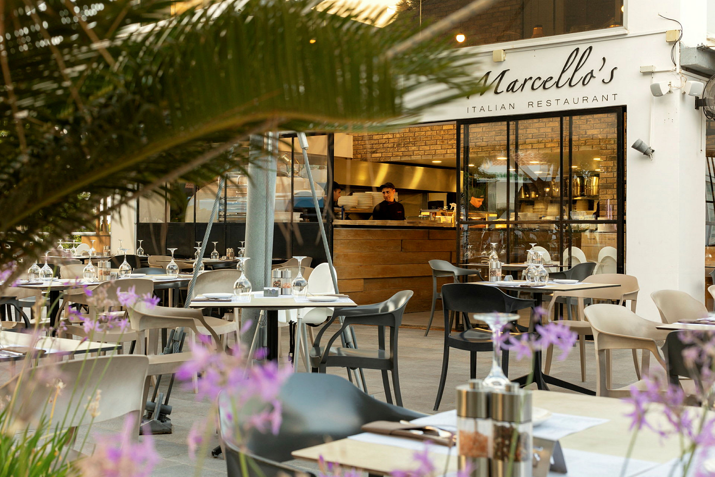 Uteservering med bord og stoler foran en italiensk restaurant med glassvegger og en tre serveringsdisk. (AI-generert tekst)