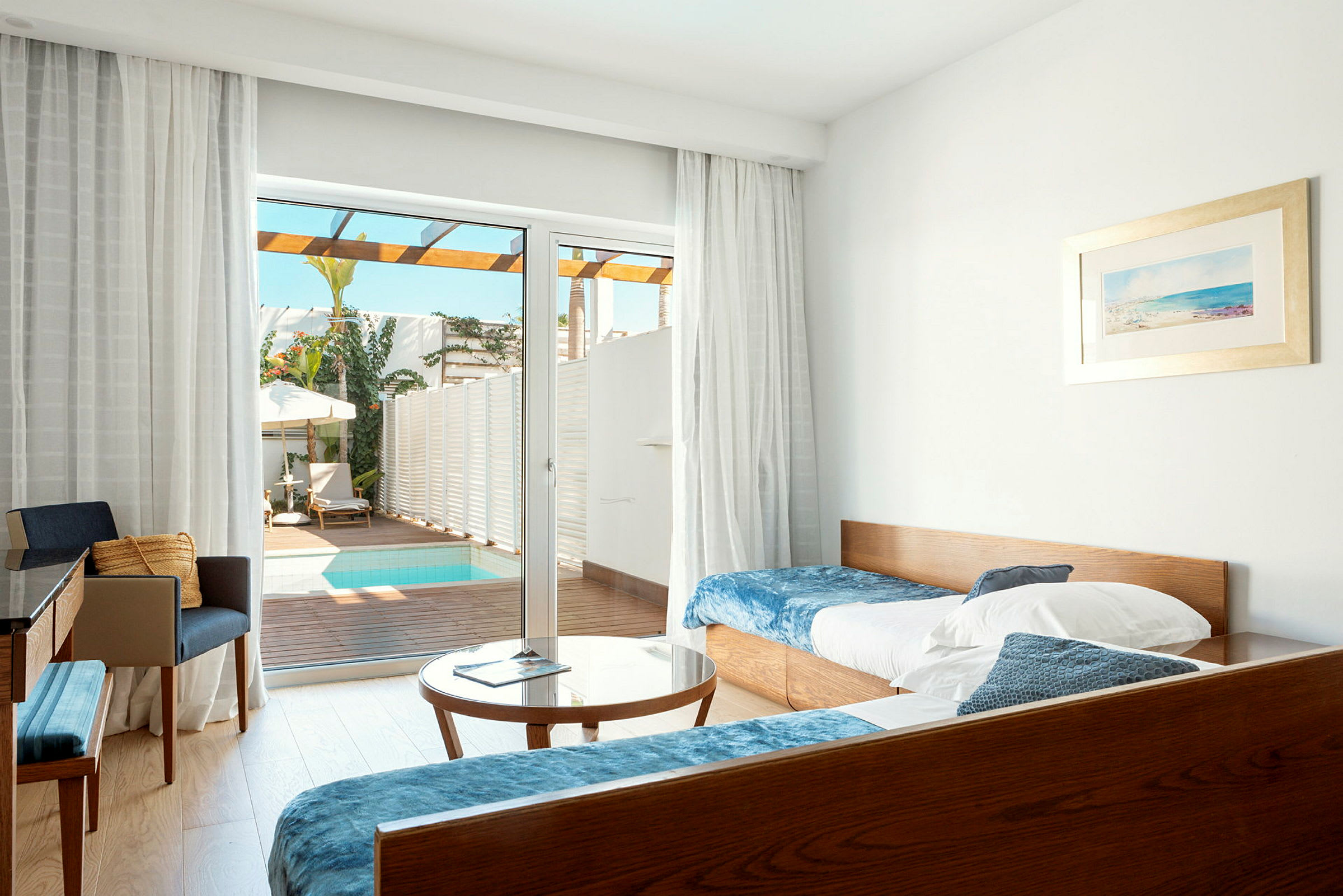 Et hotellrom med senger, bord og stoler, samt en glassdør som vender ut mot en terrasse med et lite basseng. (AI-generert tekst)