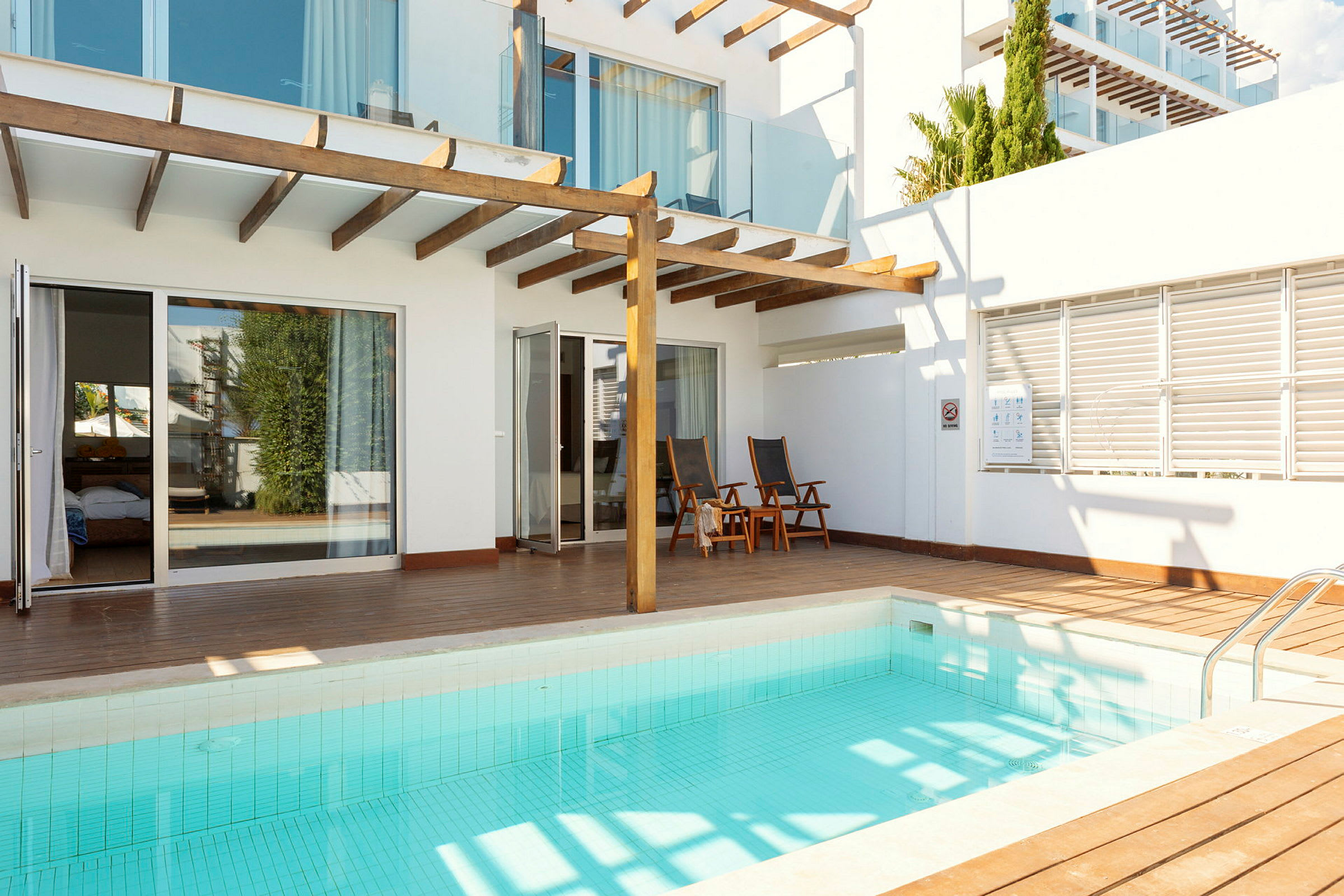 Et mindre svømmebasseng med en treterrasse og moderne hvite bygninger i bakgrunnen. (AI-generert tekst)