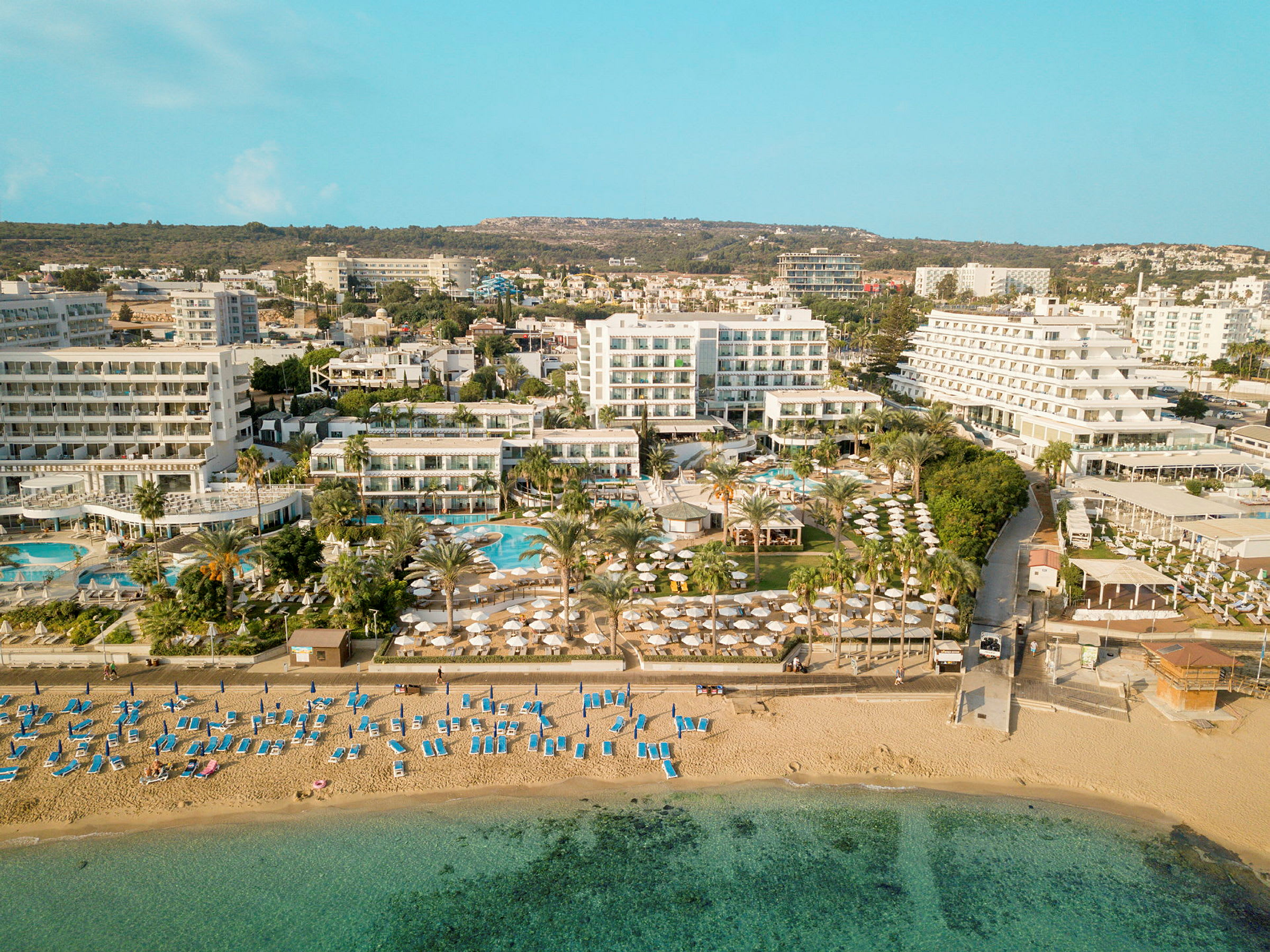 Luftfoto som viser en strand med solsenger og parasoller, samt hotellbygninger og bassengområde i bakgrunnen. (AI-generert tekst)