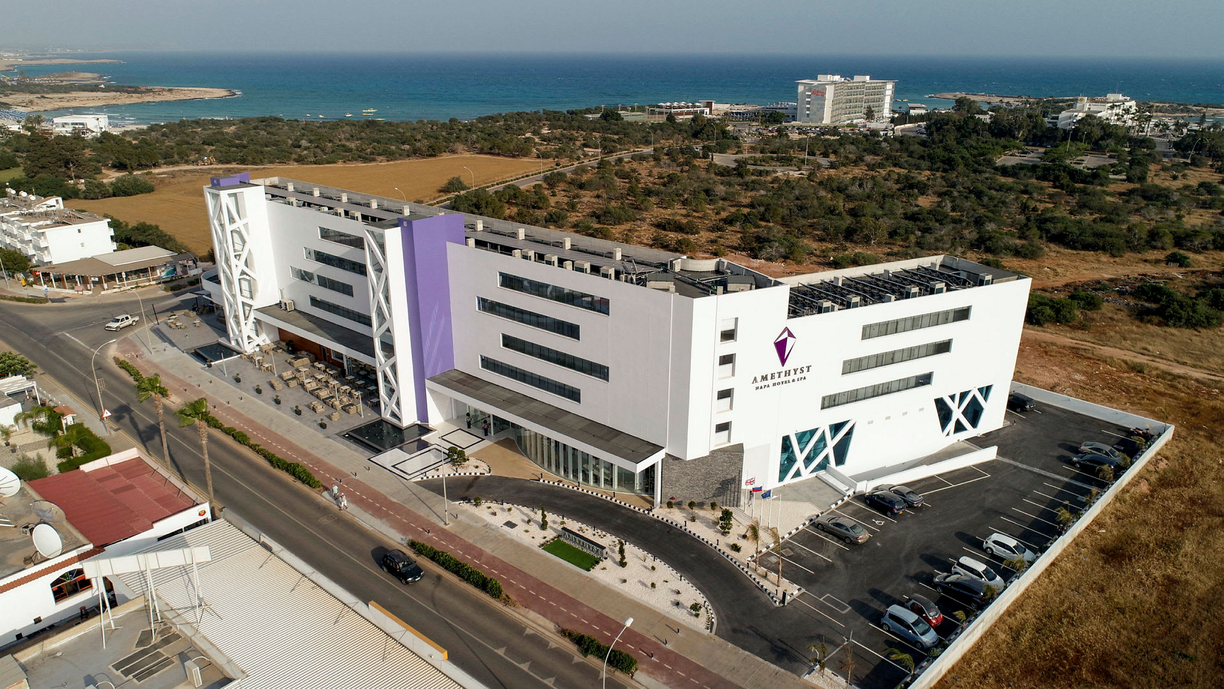 Hotellbygning med bassengområde og parkering, plassert nær kystlinjen og omgitt av grøntområder. (AI-generert tekst)