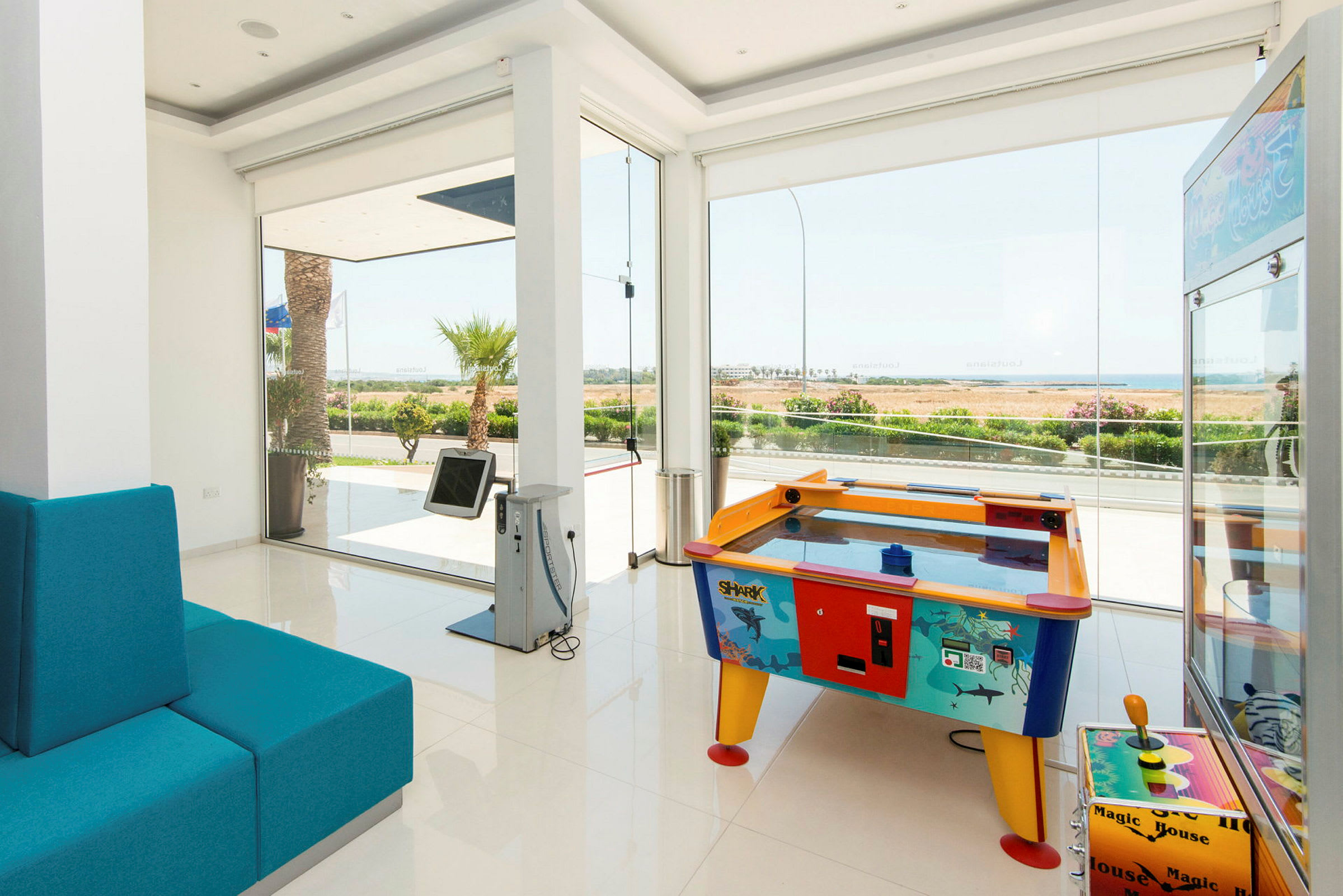 Et lyst rom med store vinduer, med en blå sofa og spill som en luftpuckmaskin samt et spill med joystick. (AI-generert tekst)