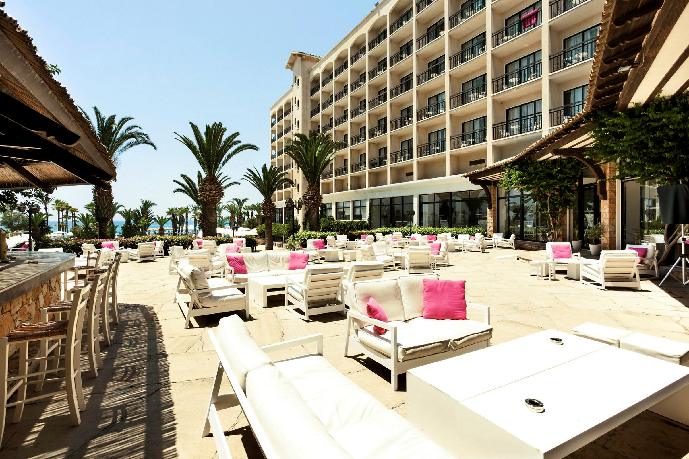 En romslig solterrasse med utemøbler i hvitt og rosa, omgitt av palmer og et stort hotell i bakgrunnen. (AI-generert tekst)